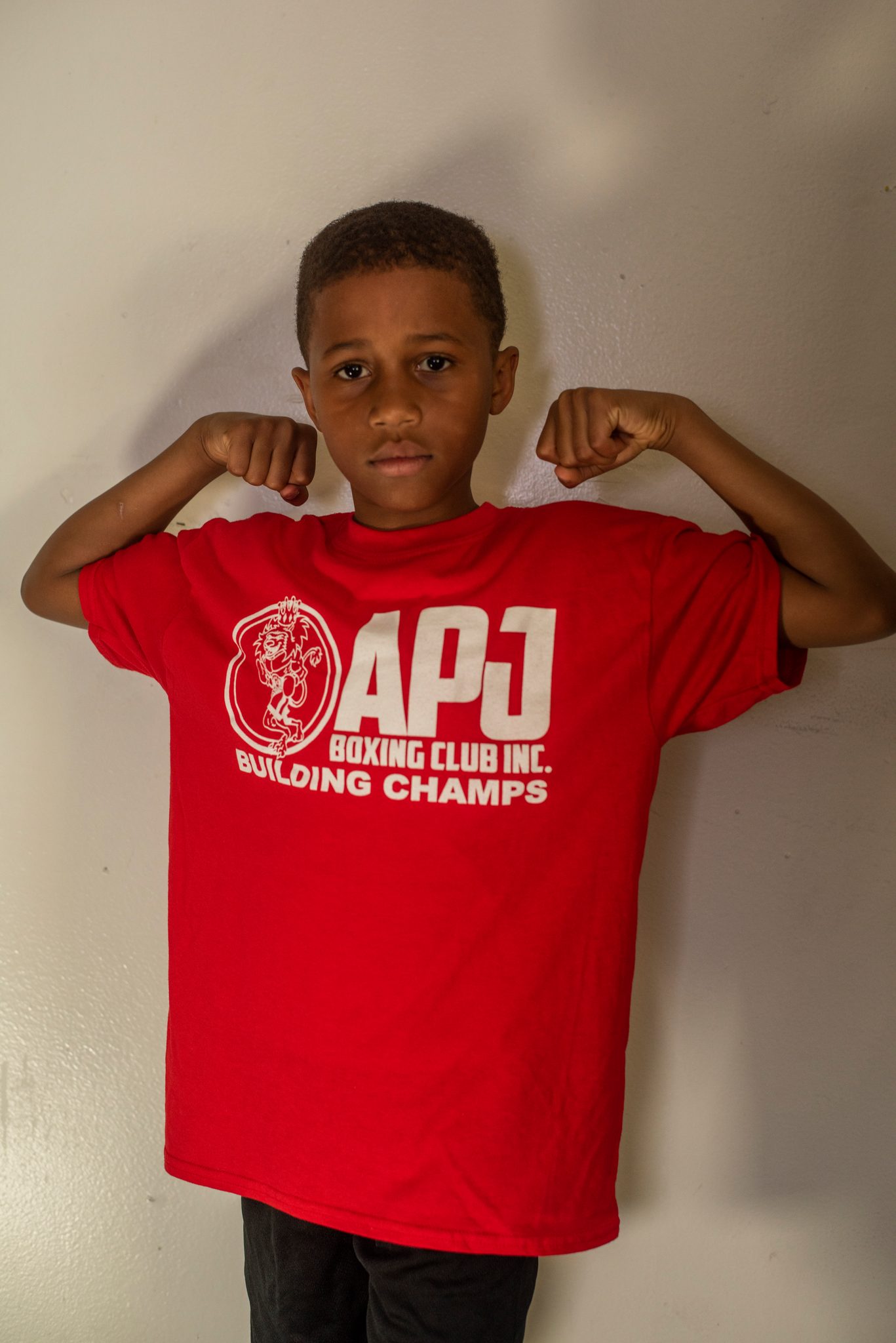 The same boy flexes both arms high, the club shirt hanging almost to his knees.