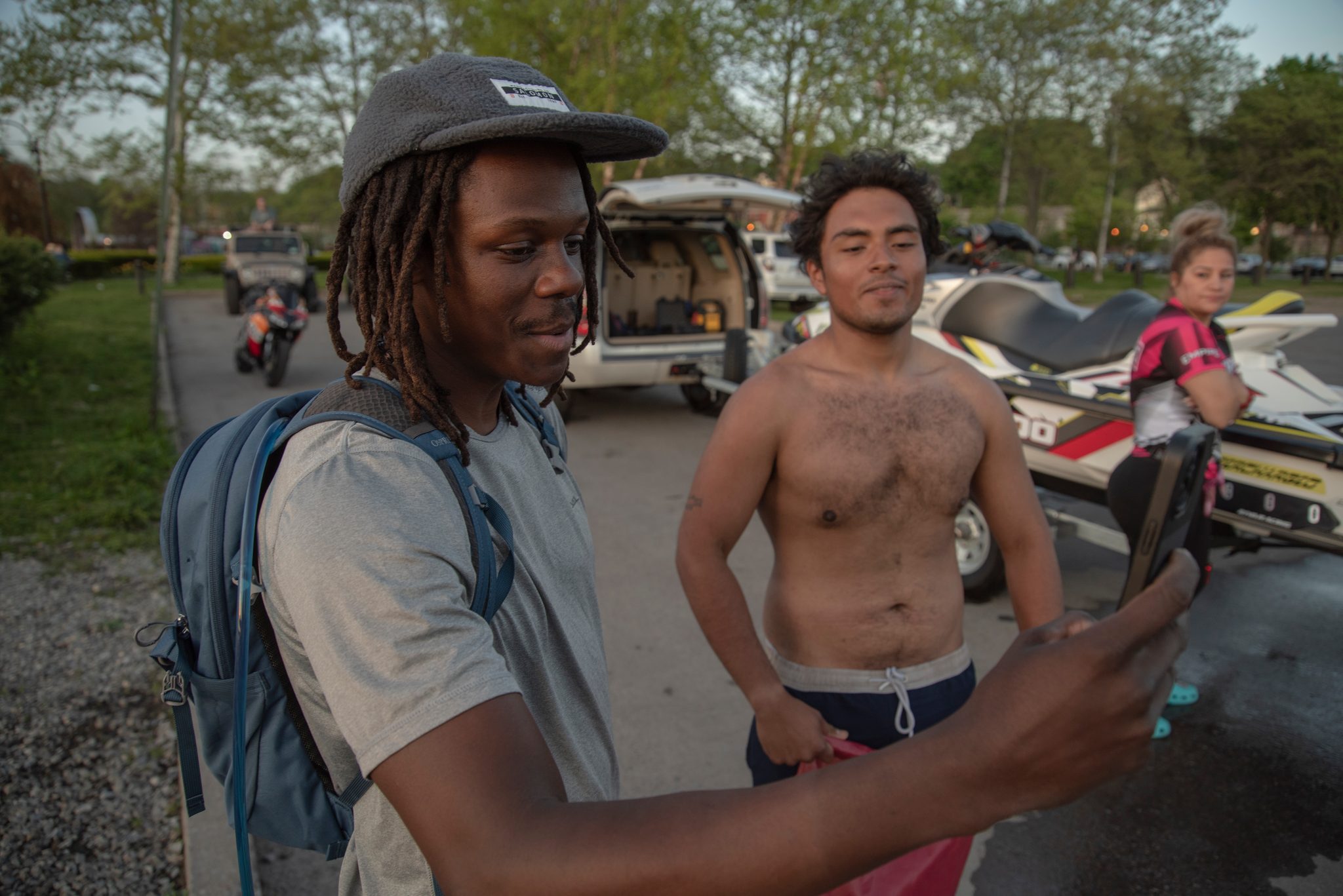 Chev holds up his phone beside a row of trailered jet skis, a companion leaning in to look.