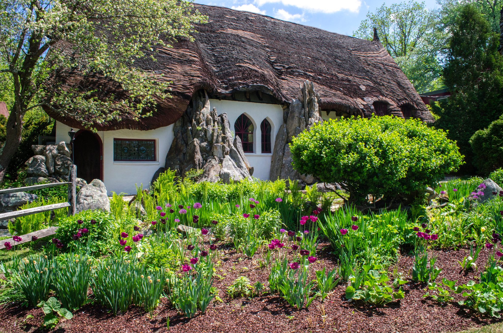 A storybook cottage with a thatched roof and stone chimneys sits behind a garden of purple allium.