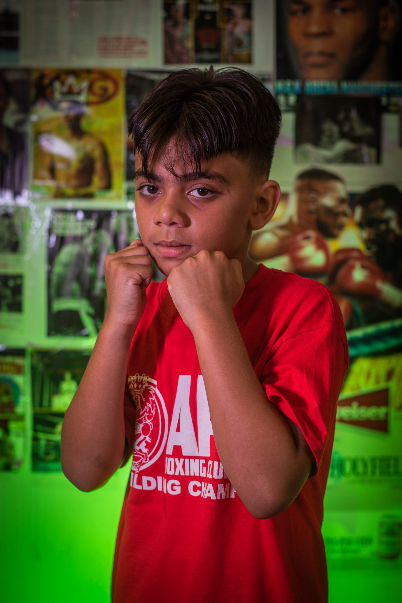 A young boxer with a sweep of dark hair holds his fists at his chin under green light.