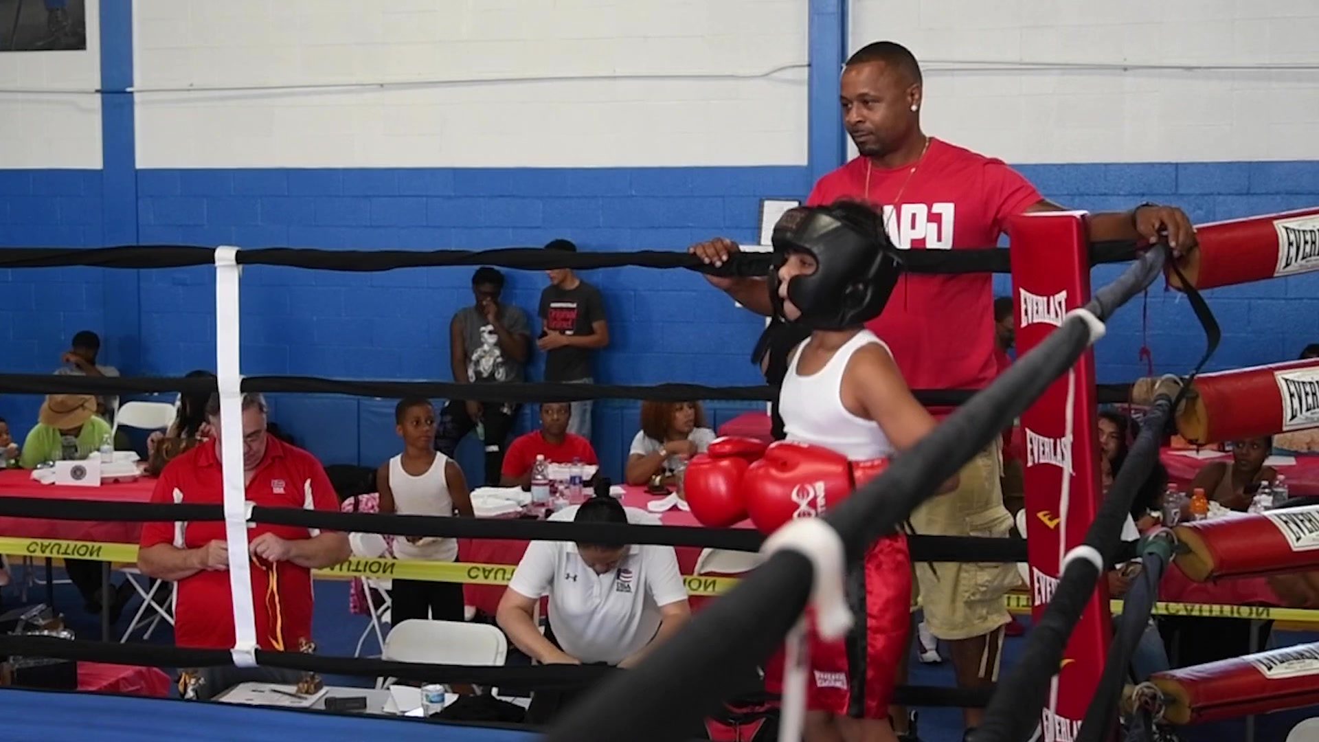 Coach Kariym stands in the corner at the Less Guns, More Boxing show, a young boxer in headgear waiting at the ropes.