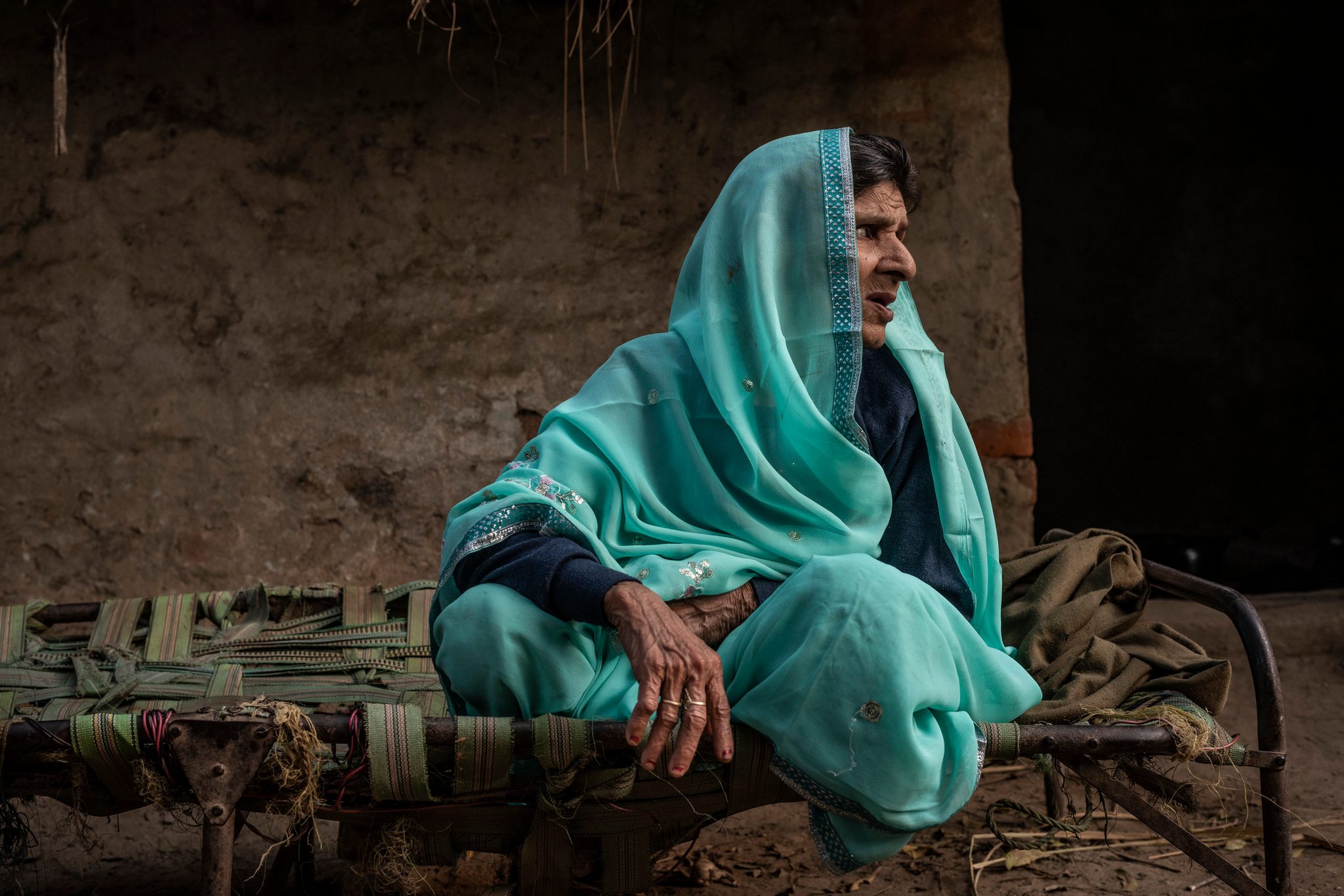 An elder in turquoise sits on her rope cot, her gaze fixed somewhere past the doorway.