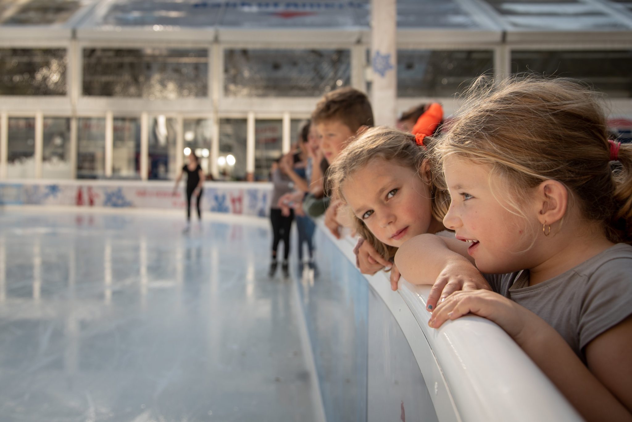 Two girls hang on the rail of the rink, chins on their arms, watching the skaters go round.