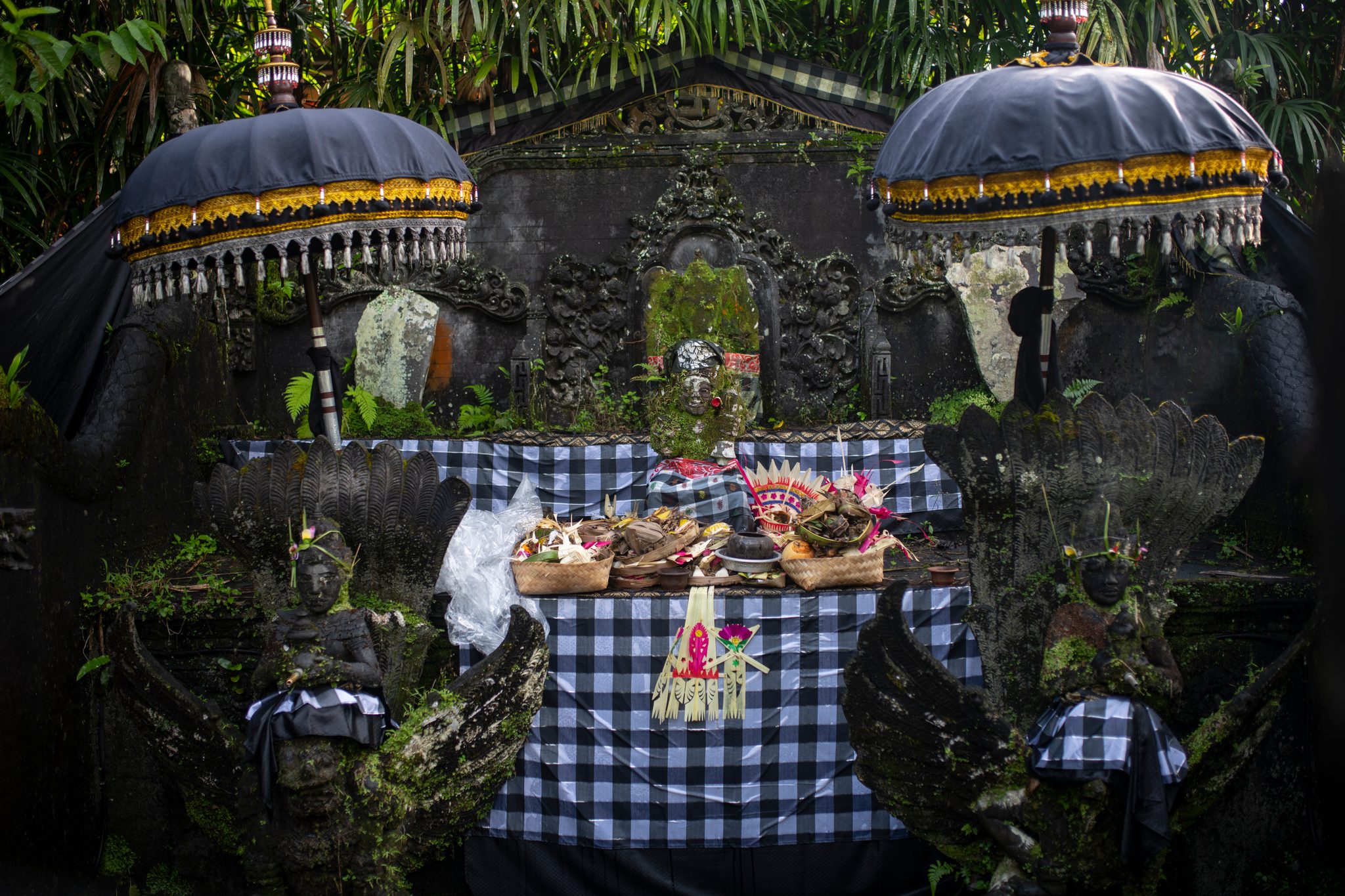 Offerings sit on the shrine beneath fringed umbrellas, the stone wrapped in black and white checked cloth.