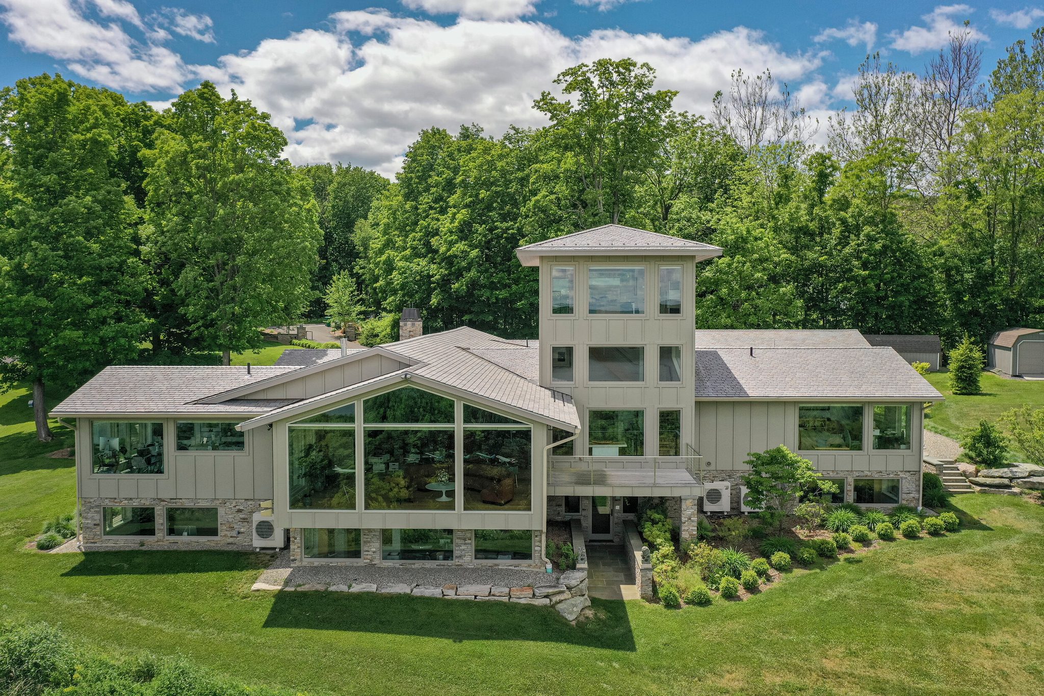 A glass-fronted contemporary with a lookout tower sits in a clearing of early June green, seen from the drone.