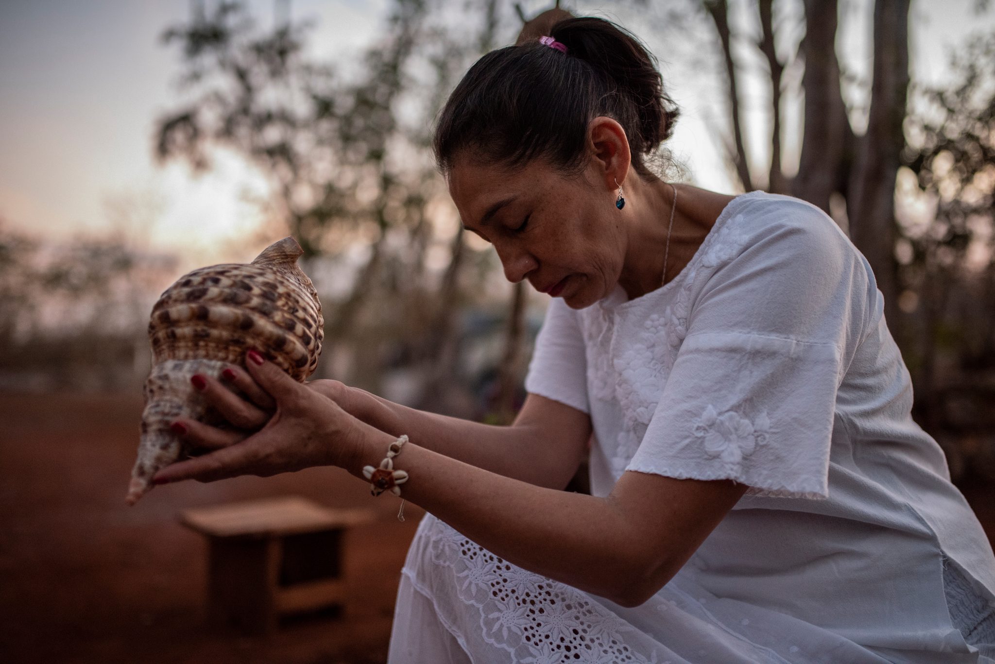 A woman in white cradles a spiral shell in both hands, eyes lowered.