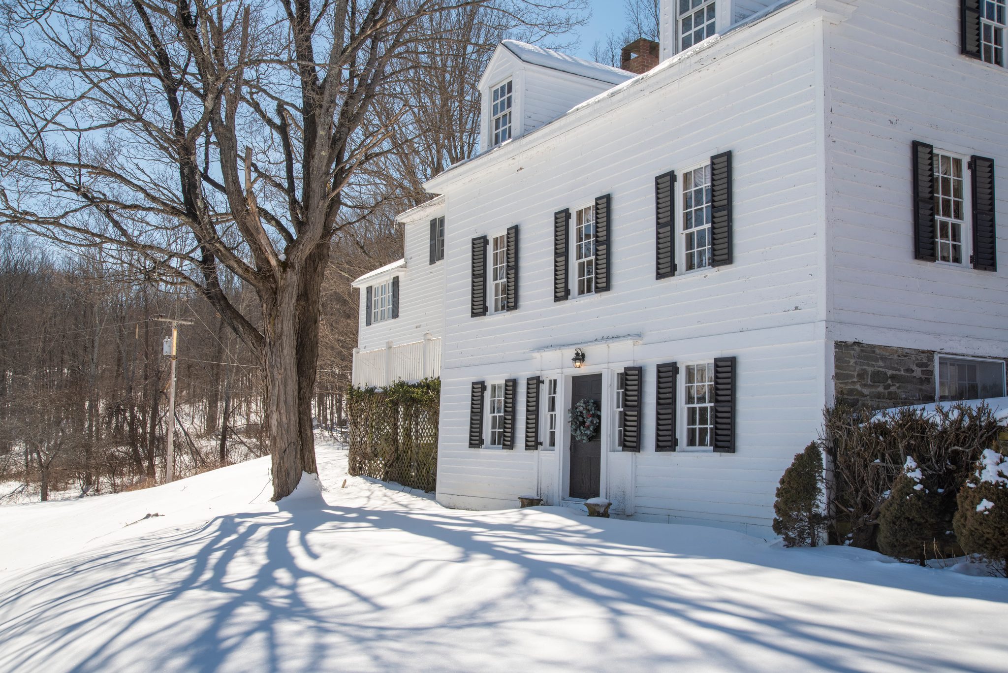 Late February sun throws bare maple shadows across deep snow in front of a white clapboard house with black shutters.