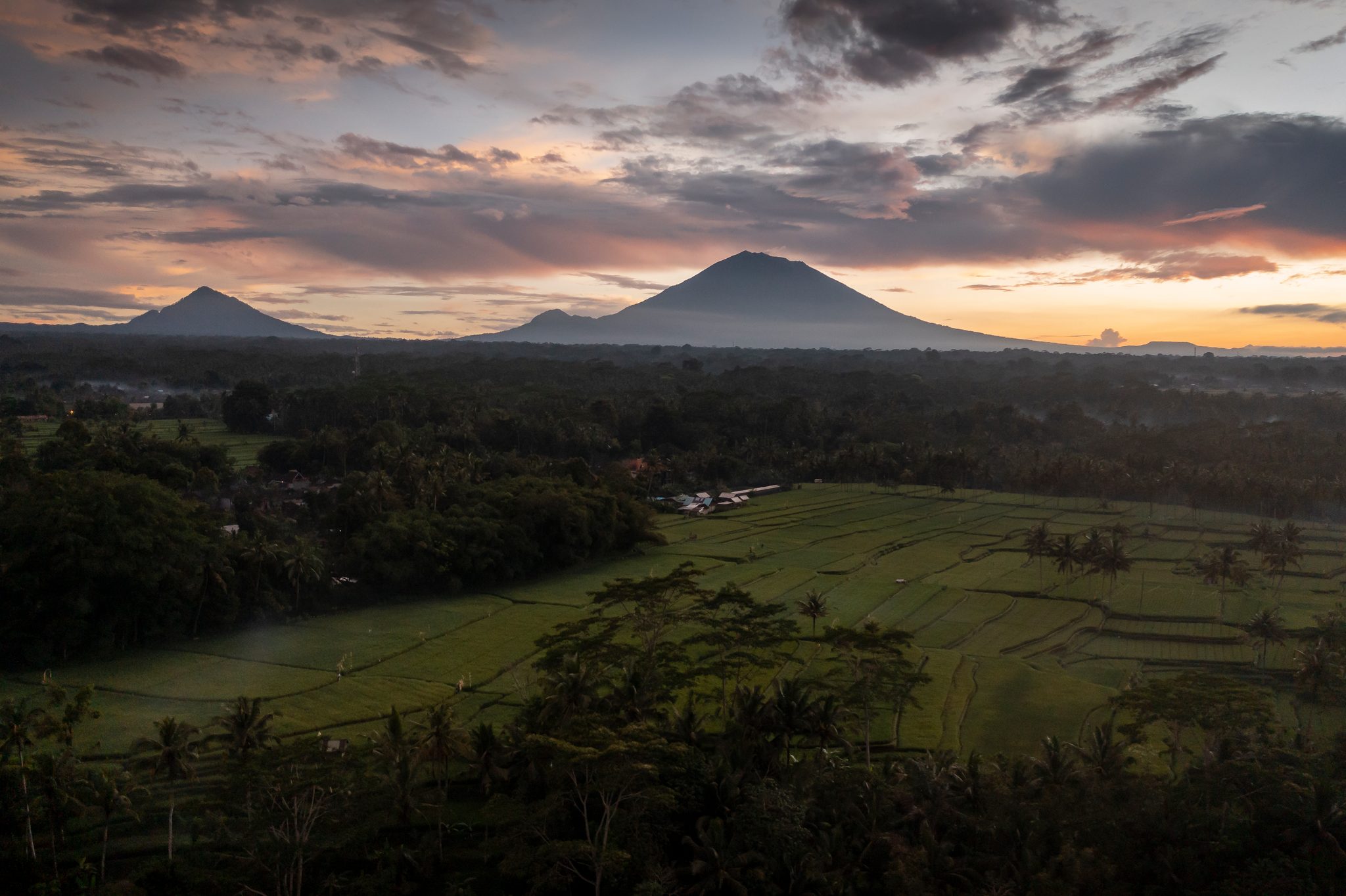 The volcano stands dark against a burning sky as the day ends over the rice fields.