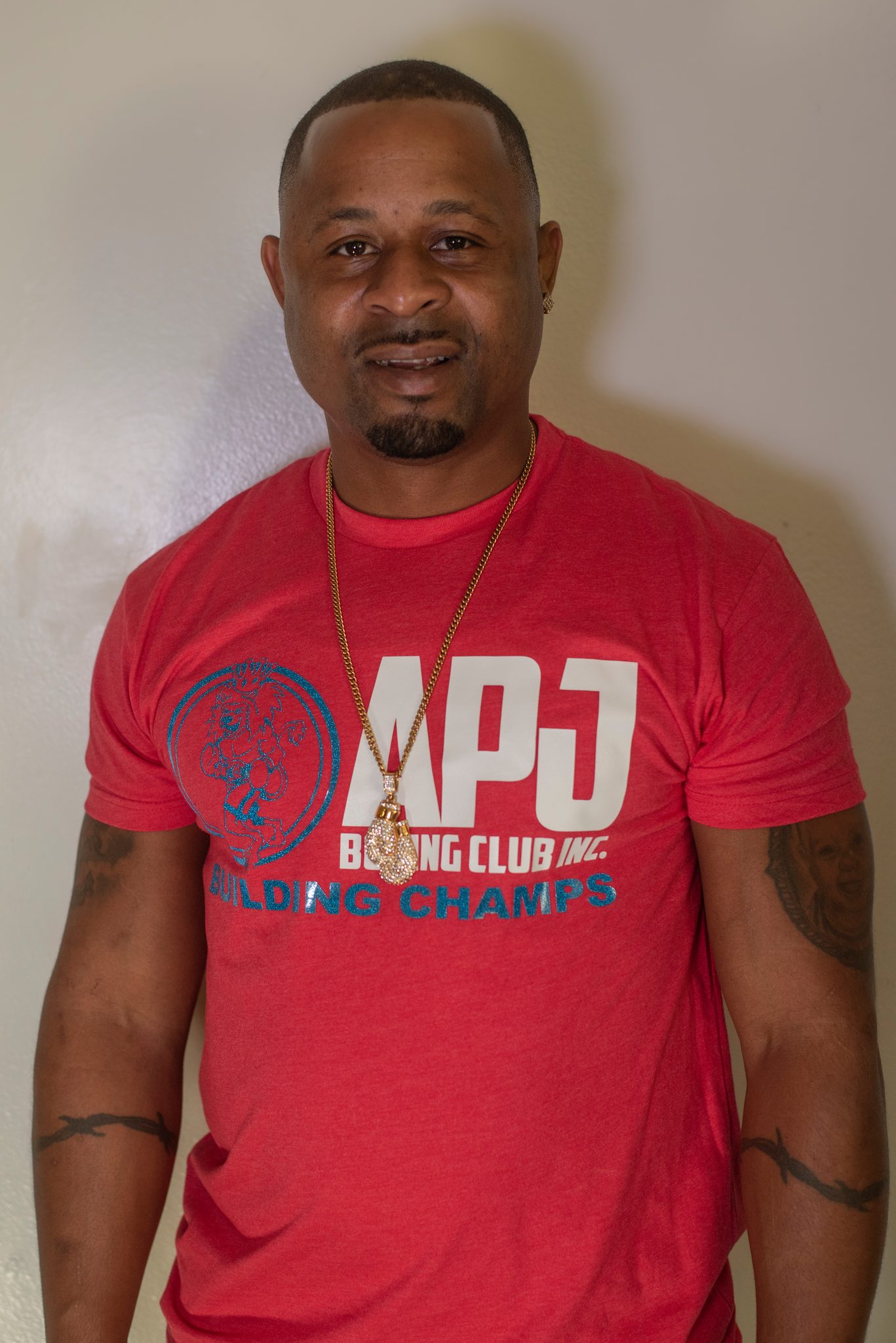 Coach Kariym relaxes into a smile in the club shirt, a gold chain and a stud earring against the white wall.