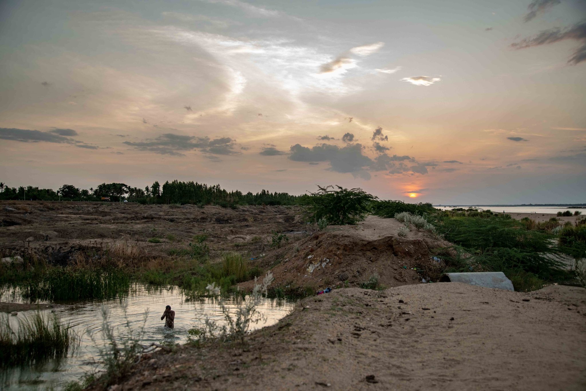 The sun sets over a riverbed of sand and shallow pools, a lone figure wading.