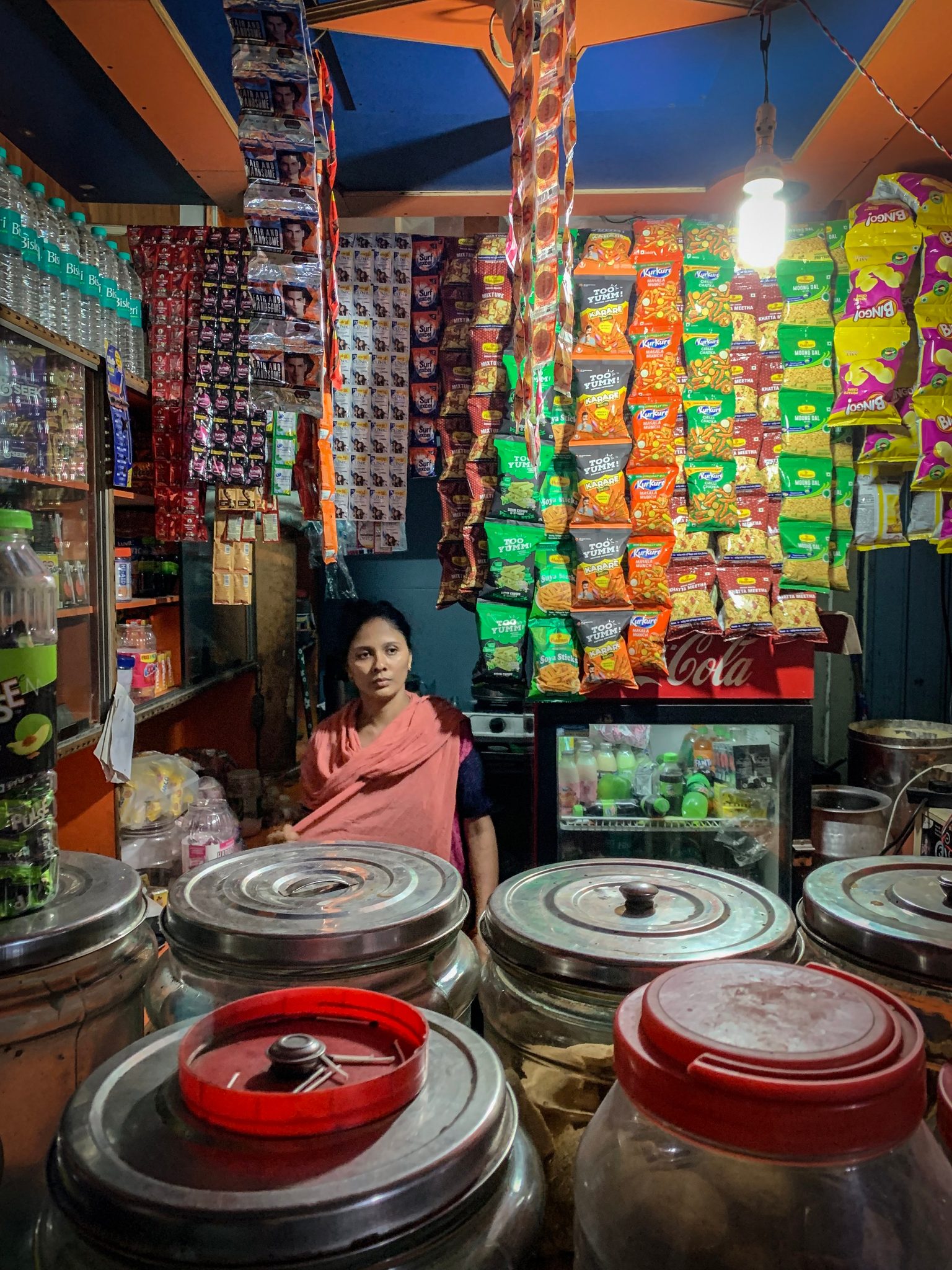 A shopkeeper sits among jars and hanging snack packets in her tiny store.