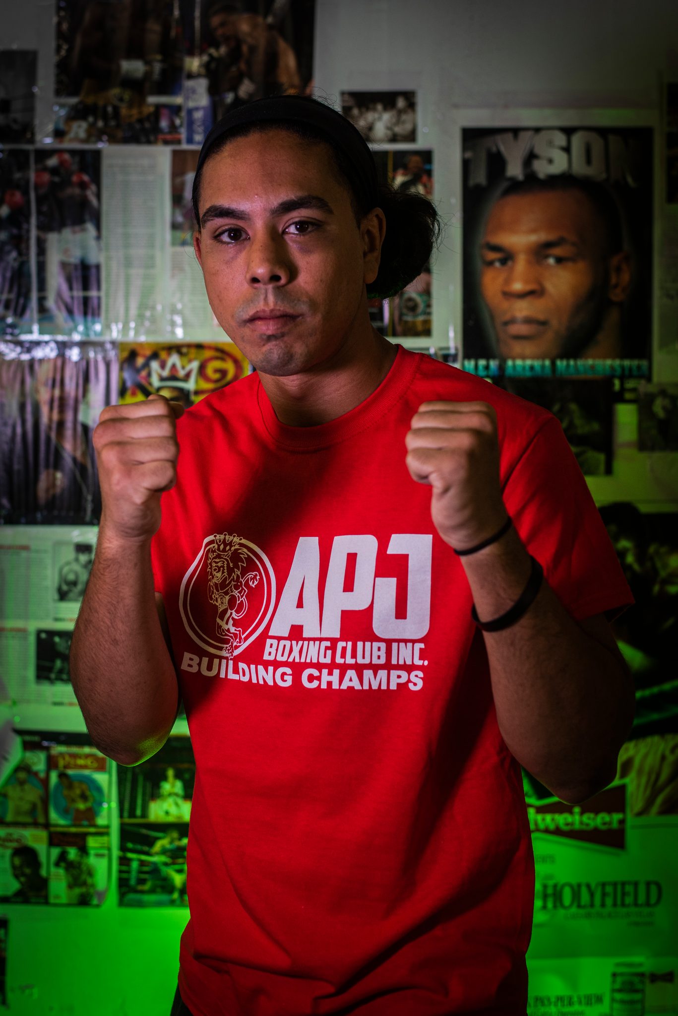 A young man with a headband and his hair tied back holds his guard up in front of a Tyson poster.