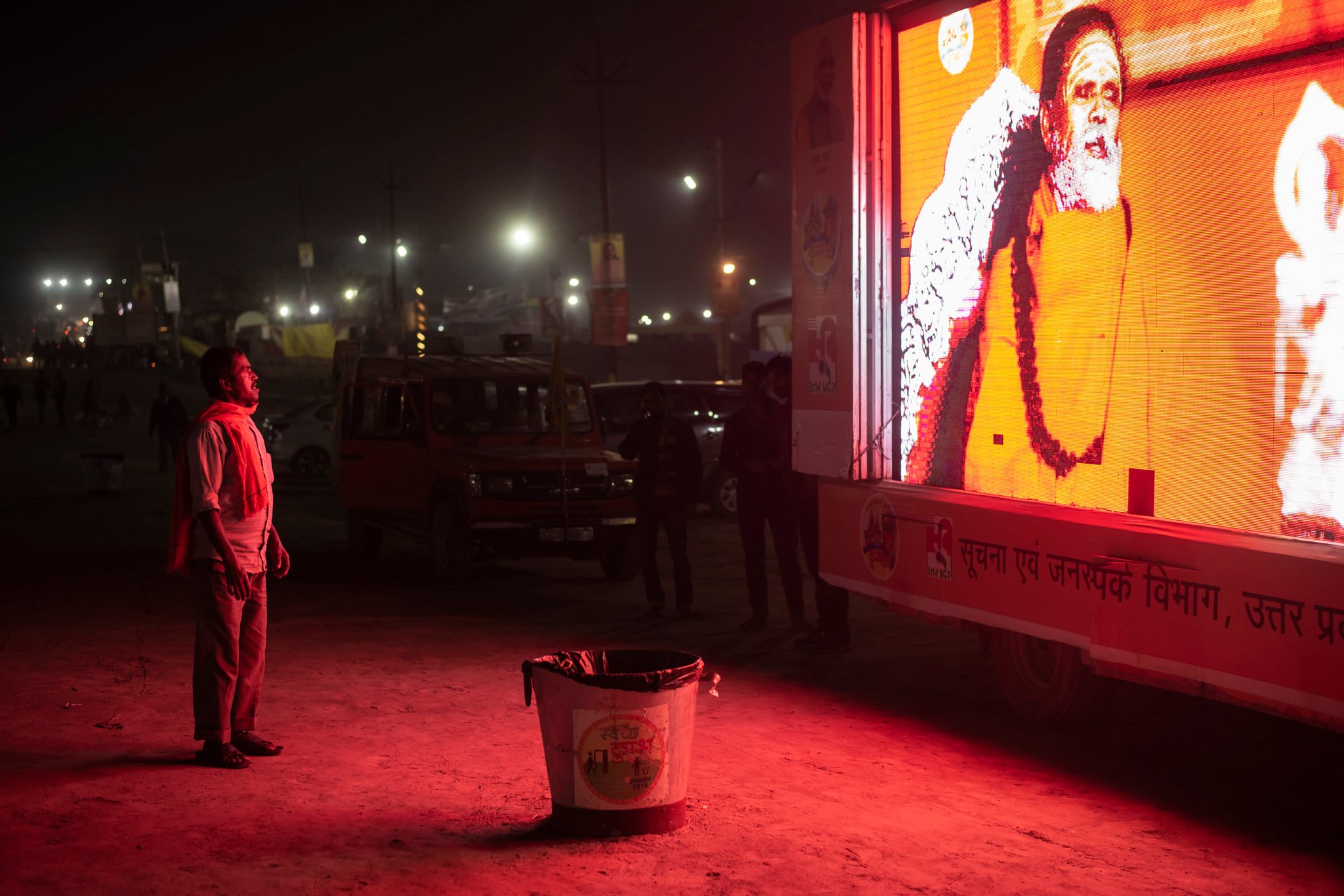 A man stands, bathed in crimson light, downloading the teachings from the glowing roadside sadhu. Here at Kumbh consciousness moves. It is pulled and pushed. For some it shrinks and others it expands.