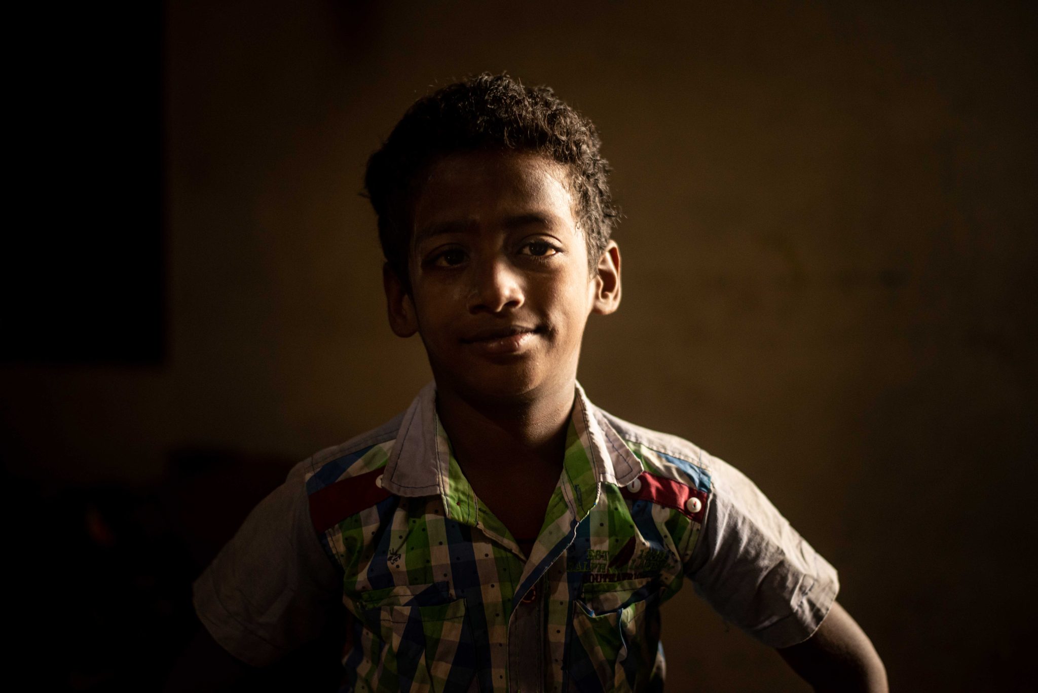 A boy smiles out of the shadows, a sliver of light across his face.