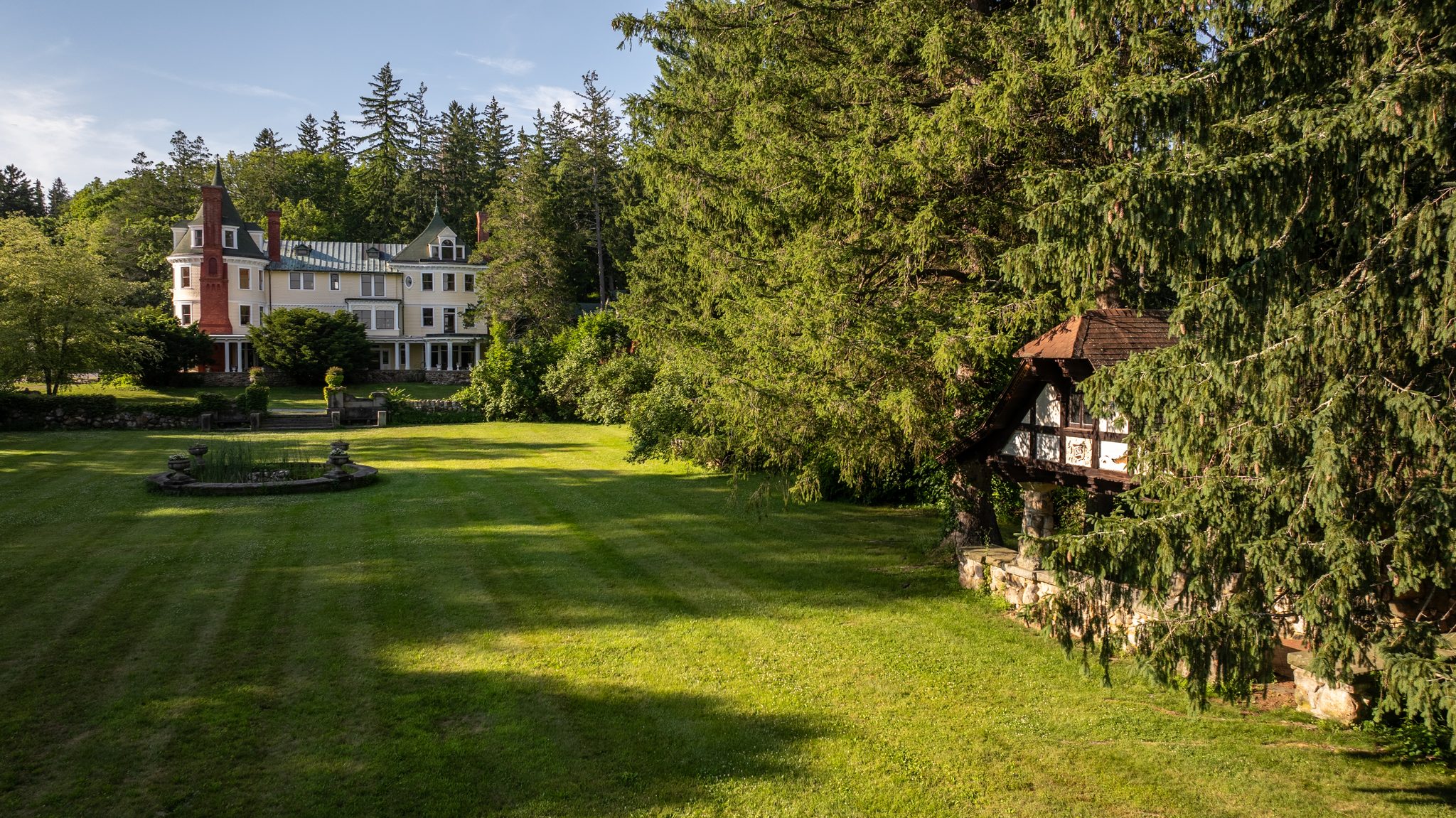 Evening shadows stretch across the lawn from a timbered pavilion toward the fountain and the main house beyond.