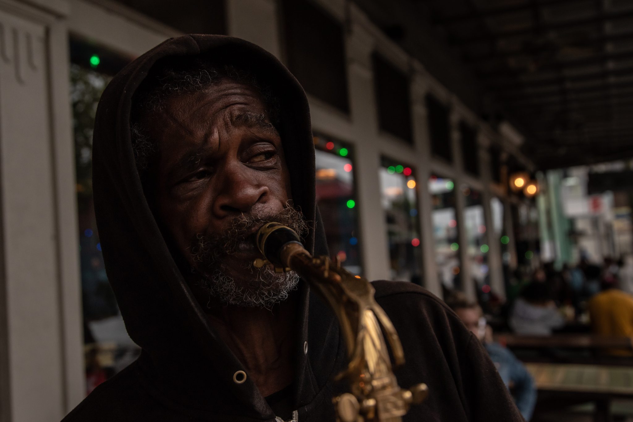 An old man in a hood plays saxophone under the gallery, eyes half closed.