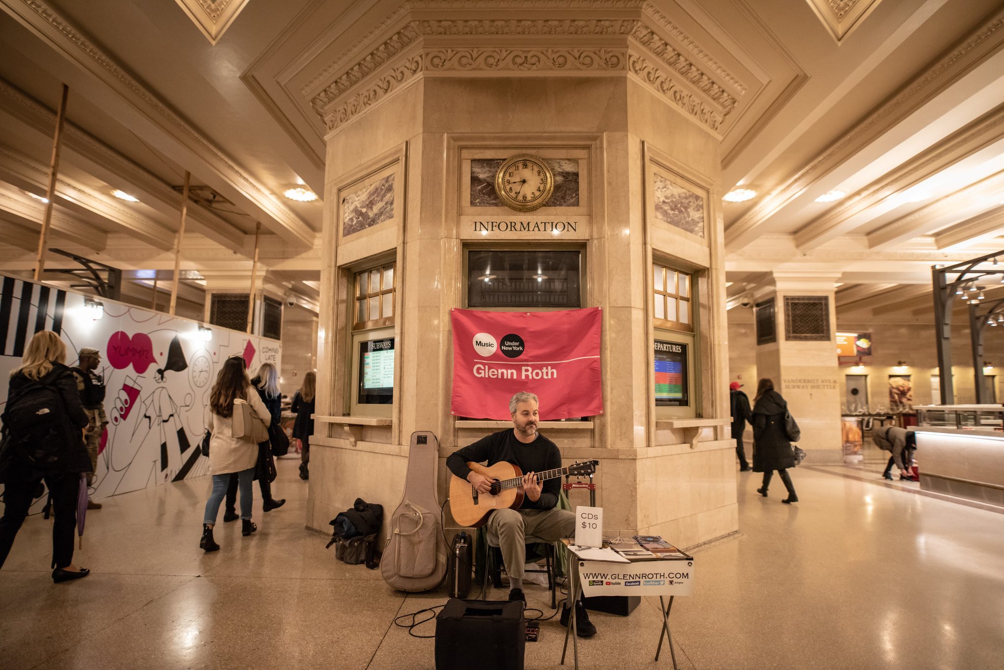 Glenn plays beneath his banner at the information booth, commuters streaming past on both sides.