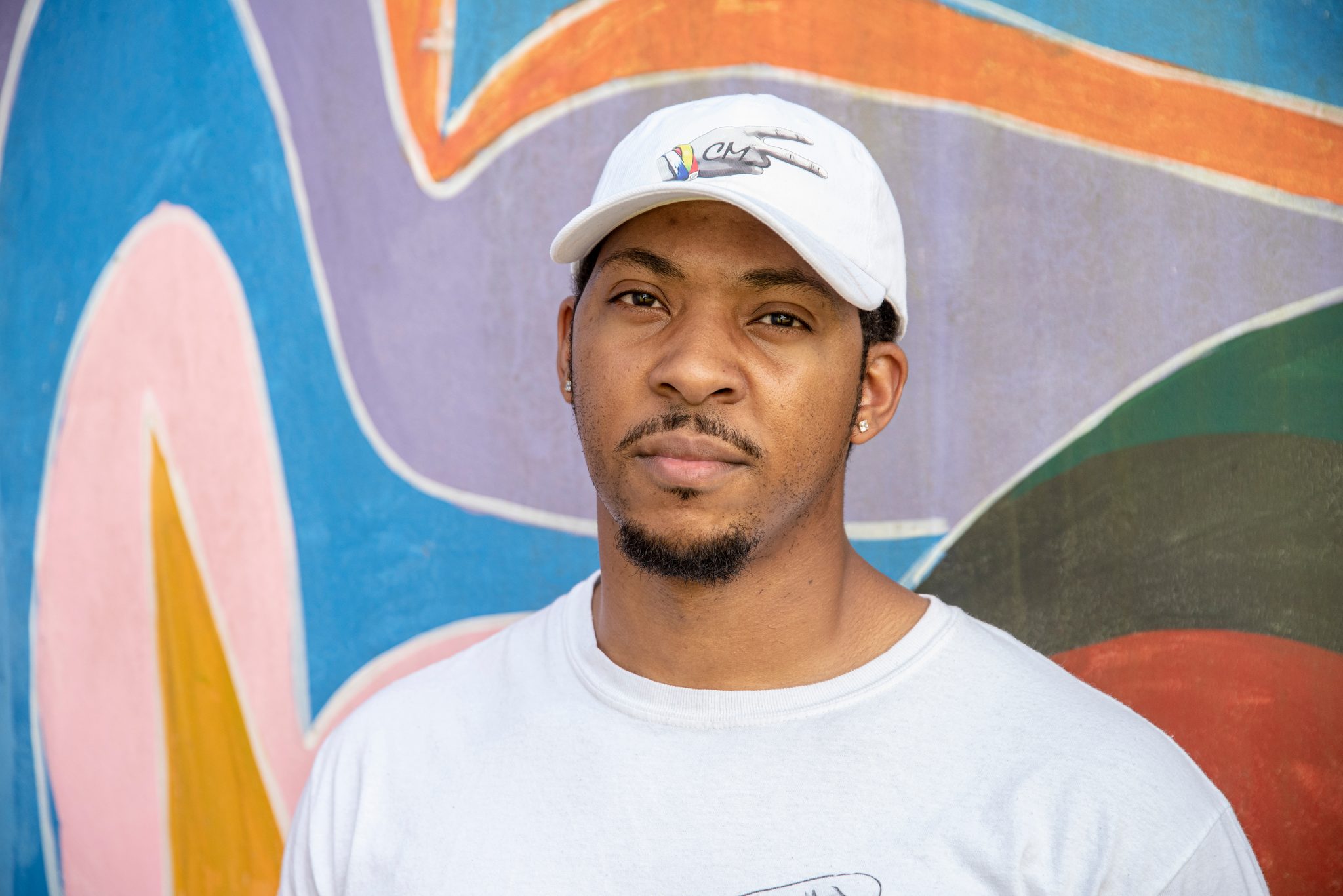 A young man in a CM2 cap stands against a mural of bright color.