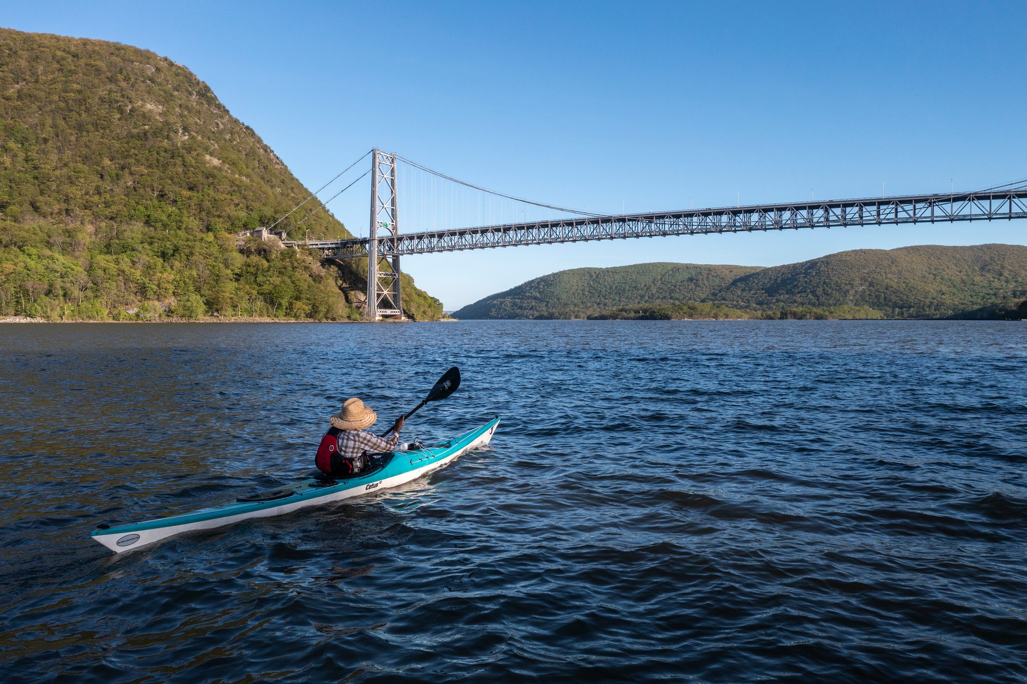From above, one teal kayak crosses open water below the Bear Mountain Bridge, the ridges green on both banks.
