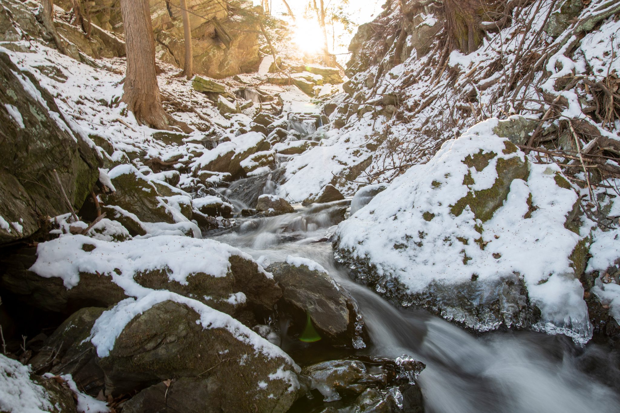 A stream runs dark between snow-covered rocks, the sun low in the gap.