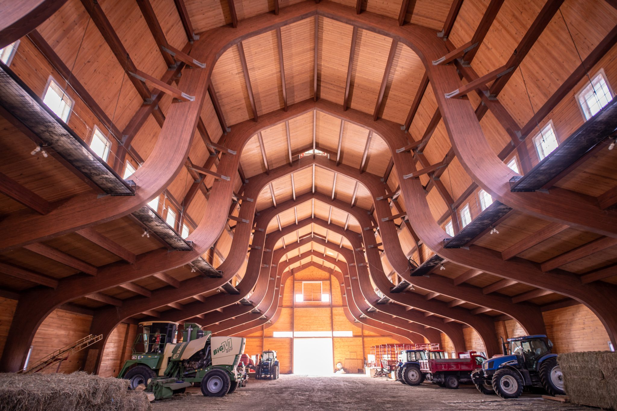 Inside a working barn in February, bent timber rafters arch overhead toward a bright doorway where the tractors wait.