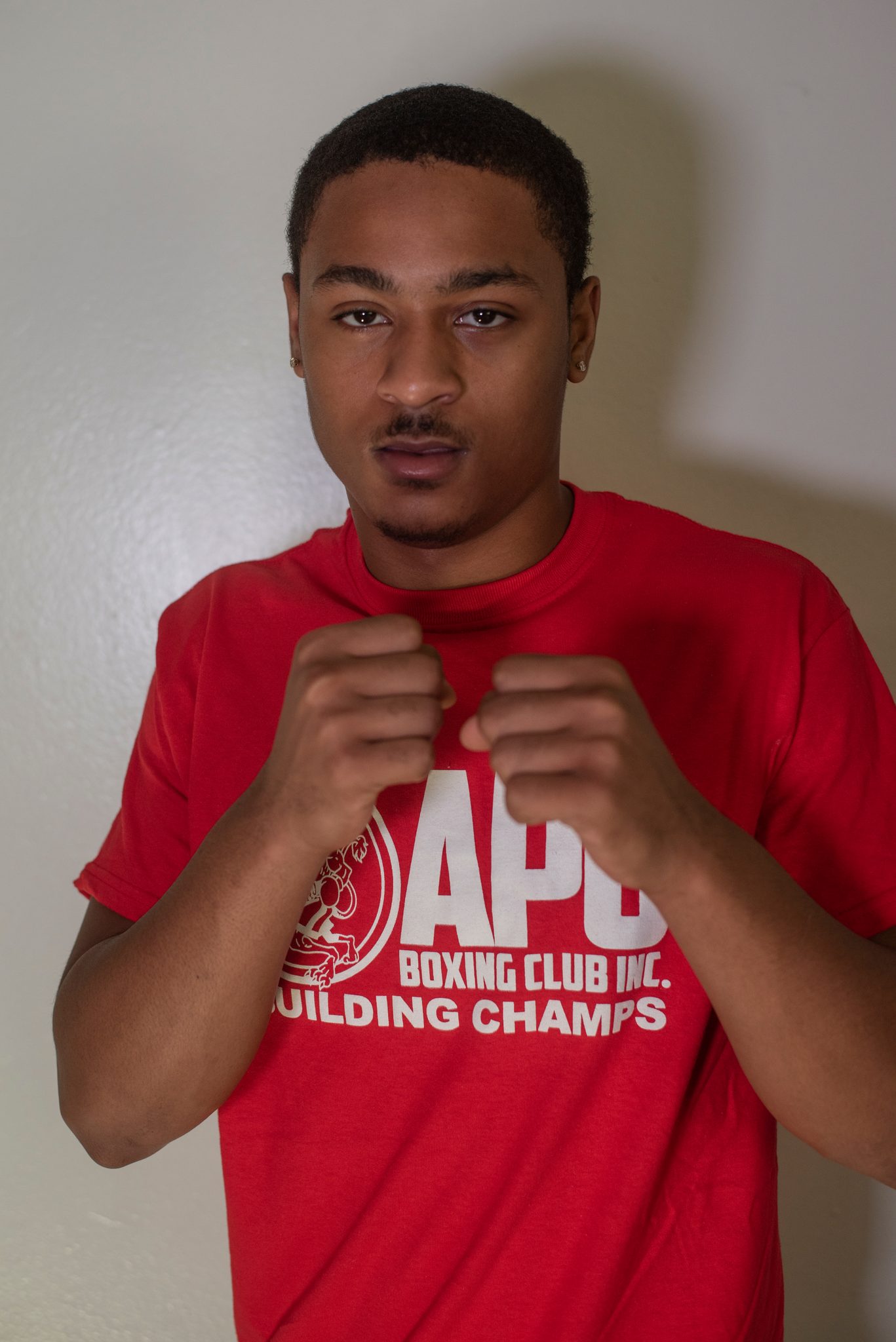 A young man with a thin mustache holds his fists at his collarbones and stares straight ahead.