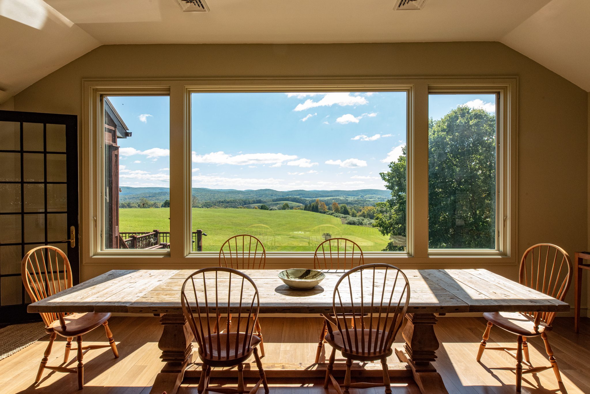 A picture window turns the farm fields into a painting above a long plank table and Windsor chairs in late September.