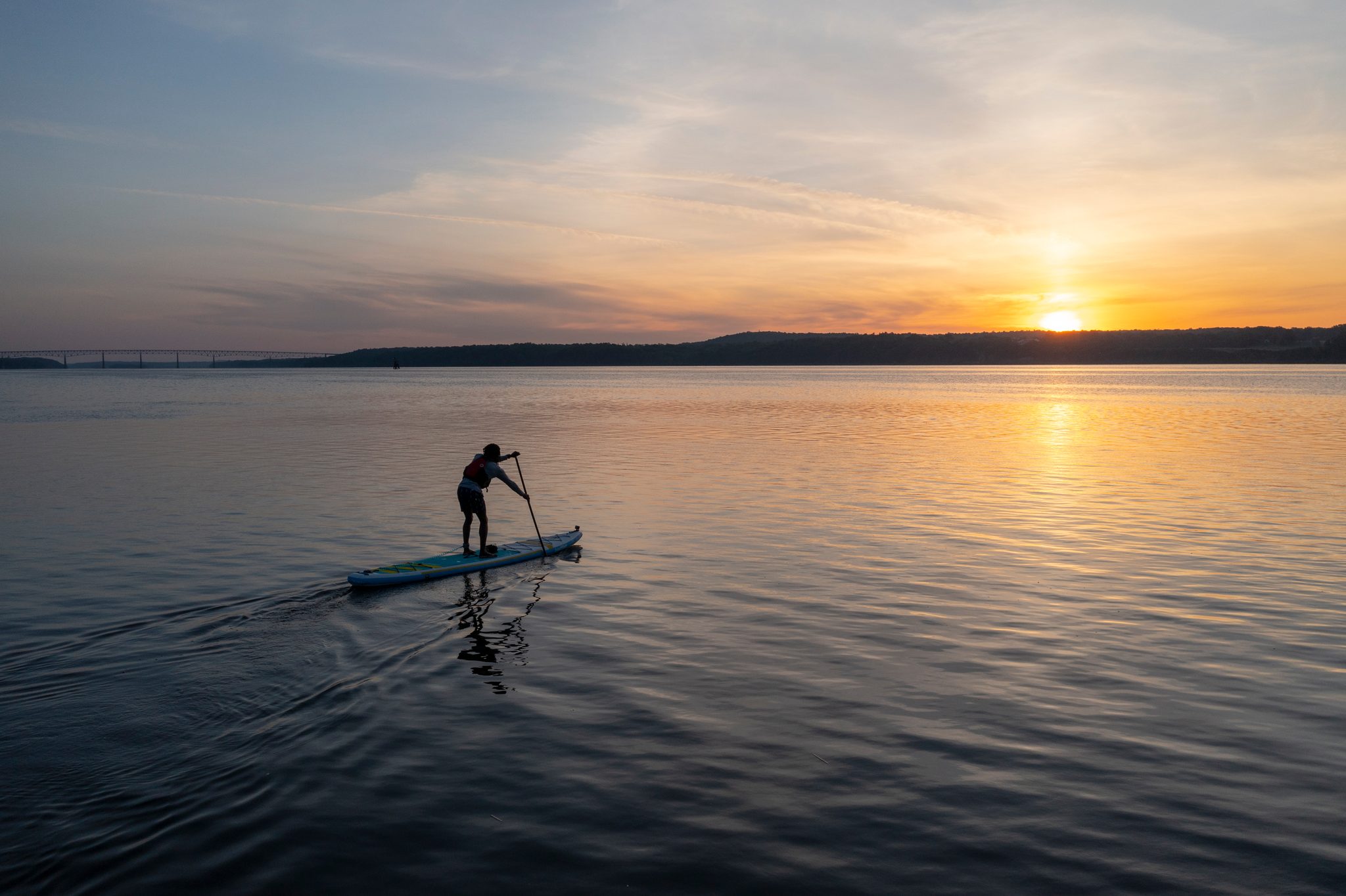 High above the river, one paddle boarder heads out toward a sun just clearing the hills.