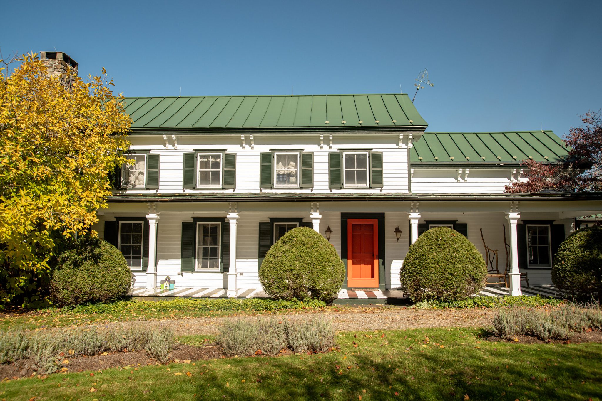 Yellow October maples frame a long white farmhouse under a green standing-seam roof and a bright red door.
