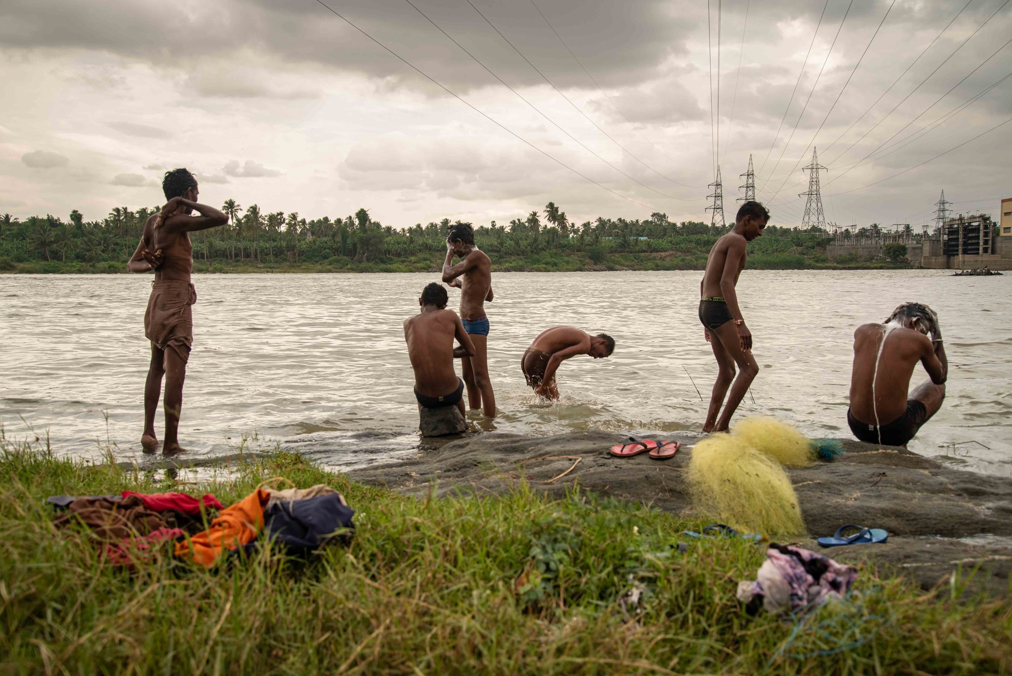 Men bathe at the river's edge beneath the power lines, a yellow net drying on the rocks.