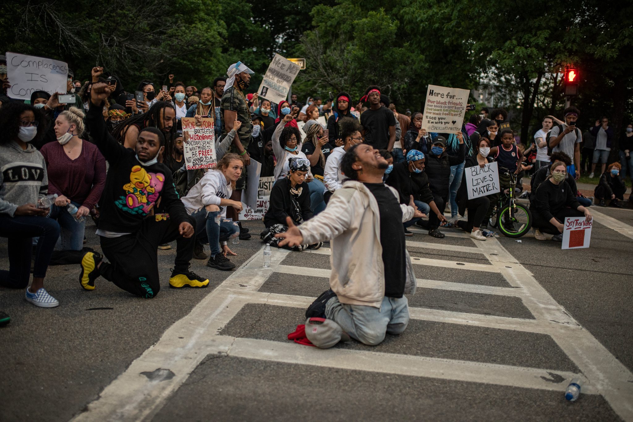A man kneels in the crosswalk, arms open and face to the sky. Around him, the crowd kneels too.