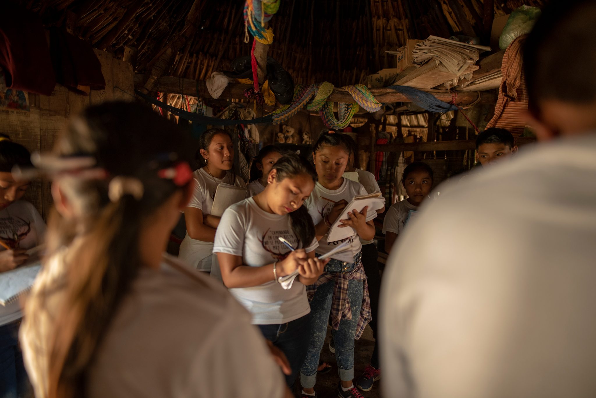 Students take notes beneath a thatched roof hung with bags and papers.