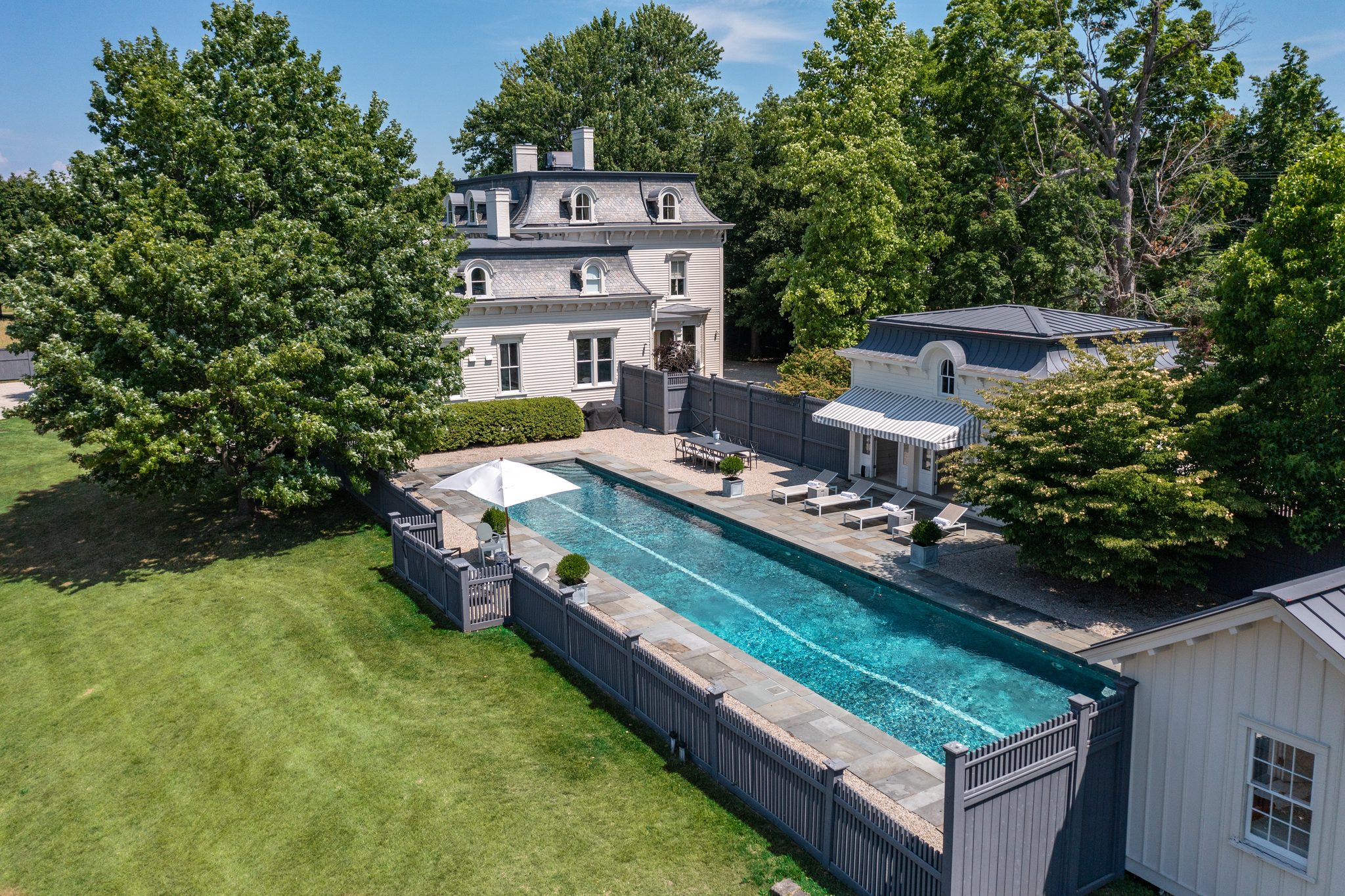 Late June from above, a mansard-roofed house and its outbuildings share a long lap pool behind a green hedge.