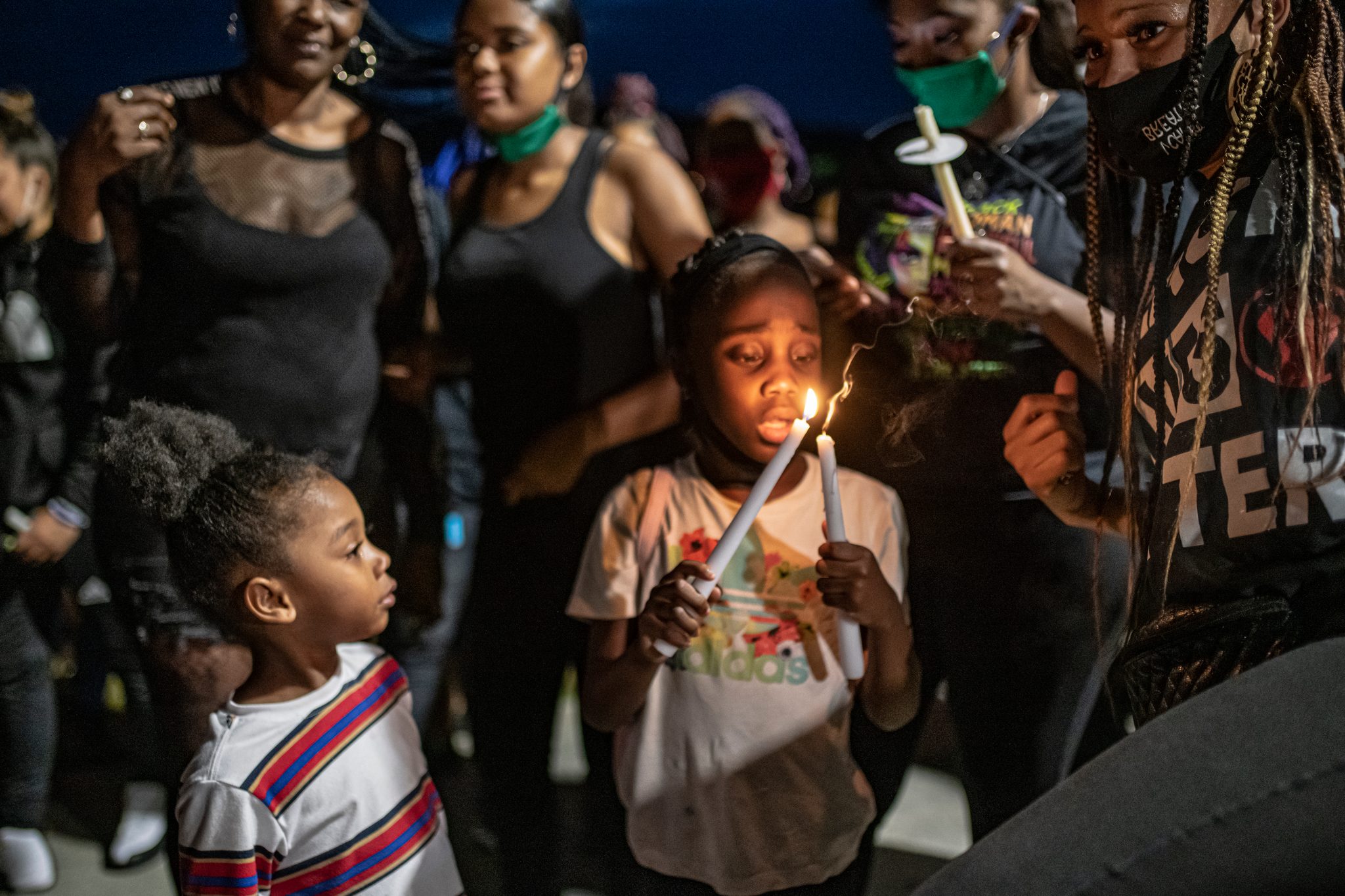 A girl cradles her candle flame. A younger one watches it, the adults standing close around them.