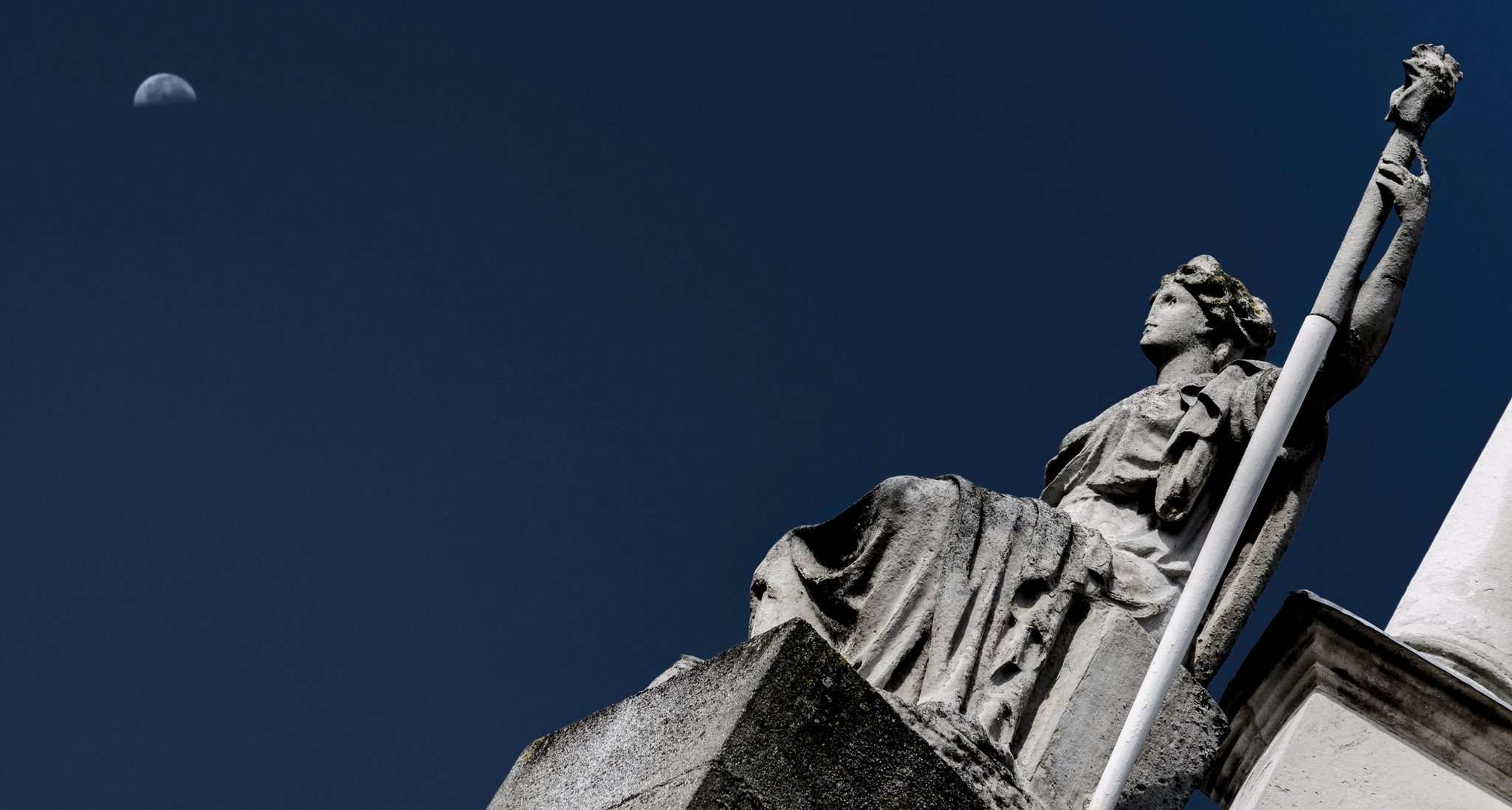 A stone statue raises her torch toward a pale half moon.