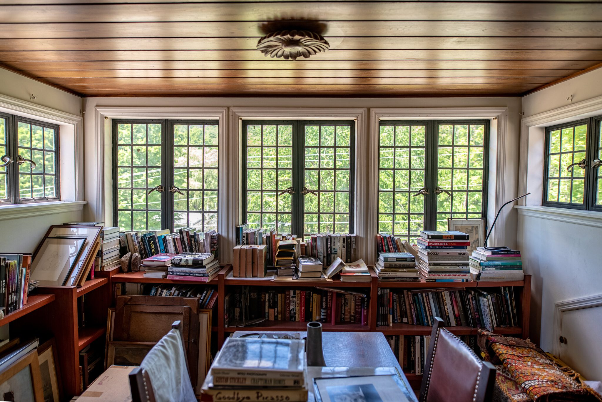 Summer green presses against steel casement windows in a book-lined sunroom under a varnished plank ceiling.