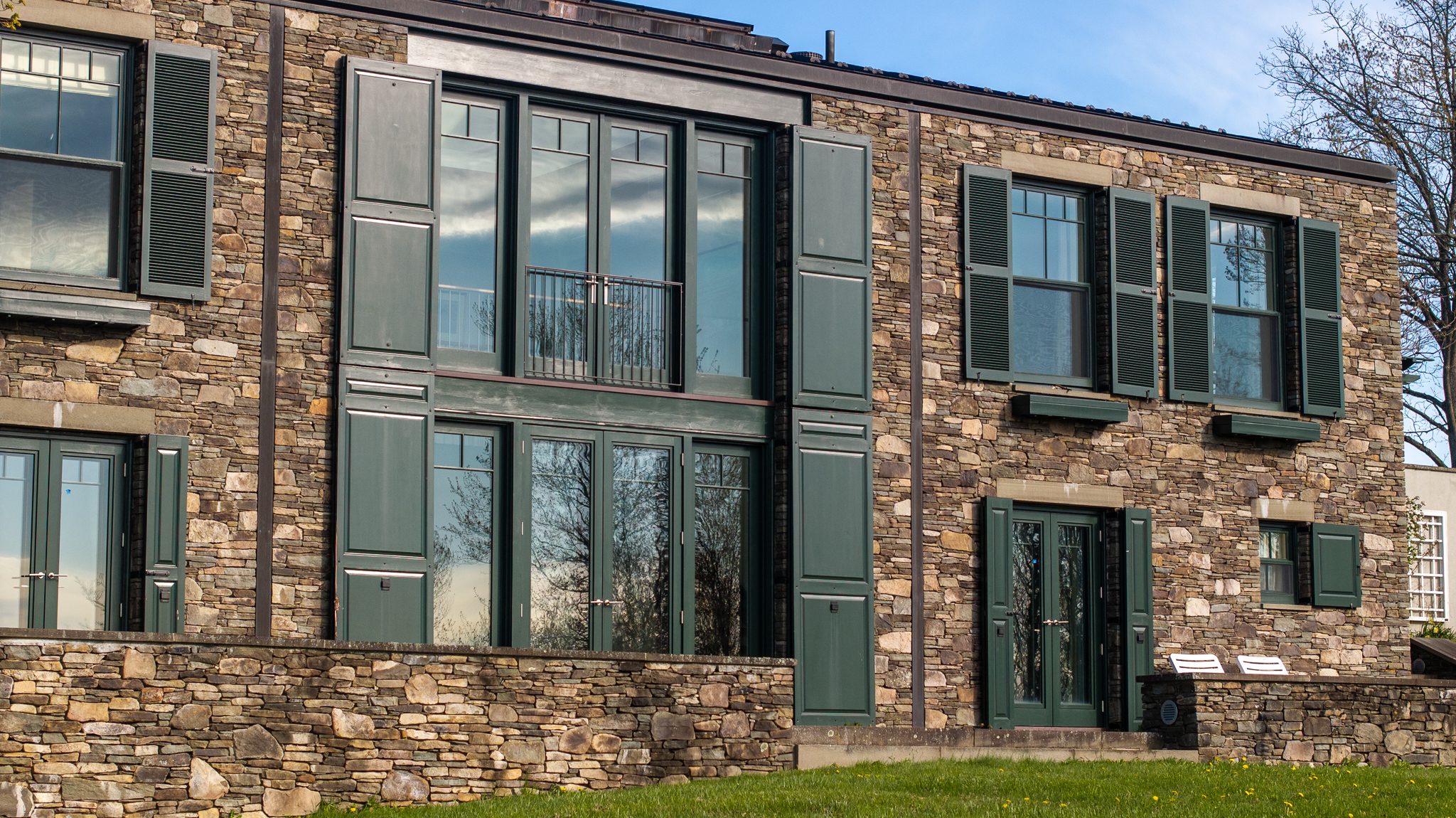 Stacked fieldstone, dark green shutters and a two-story window wall catch low late April afternoon light.
