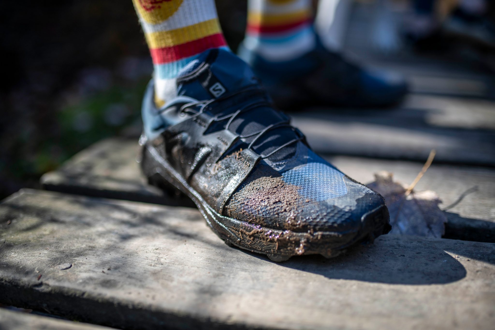 A muddy trail shoe planted on the boardwalk, striped socks above it.