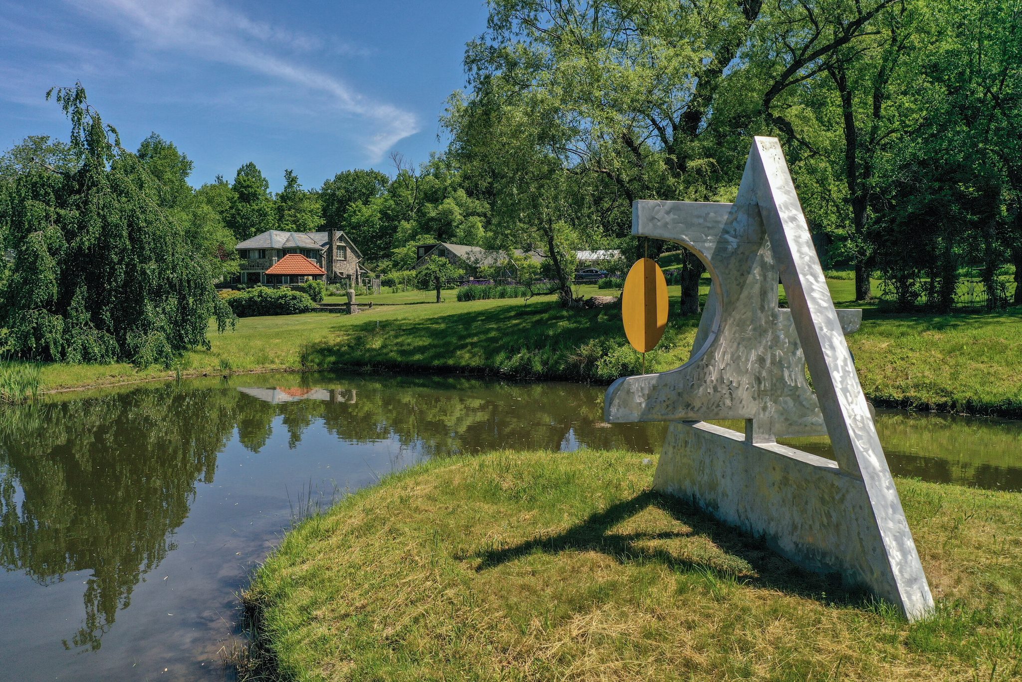 A steel sculpture stands at the edge of a still June pond, the house tucked into the trees across the water.