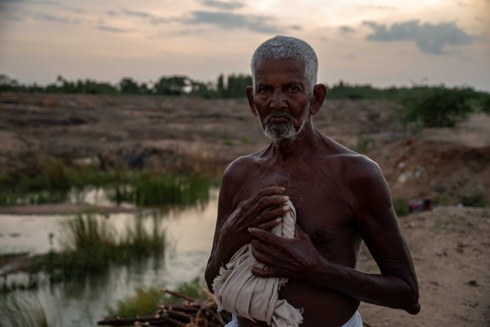 An elder holds a cloth to his chest as the sun goes down behind him.