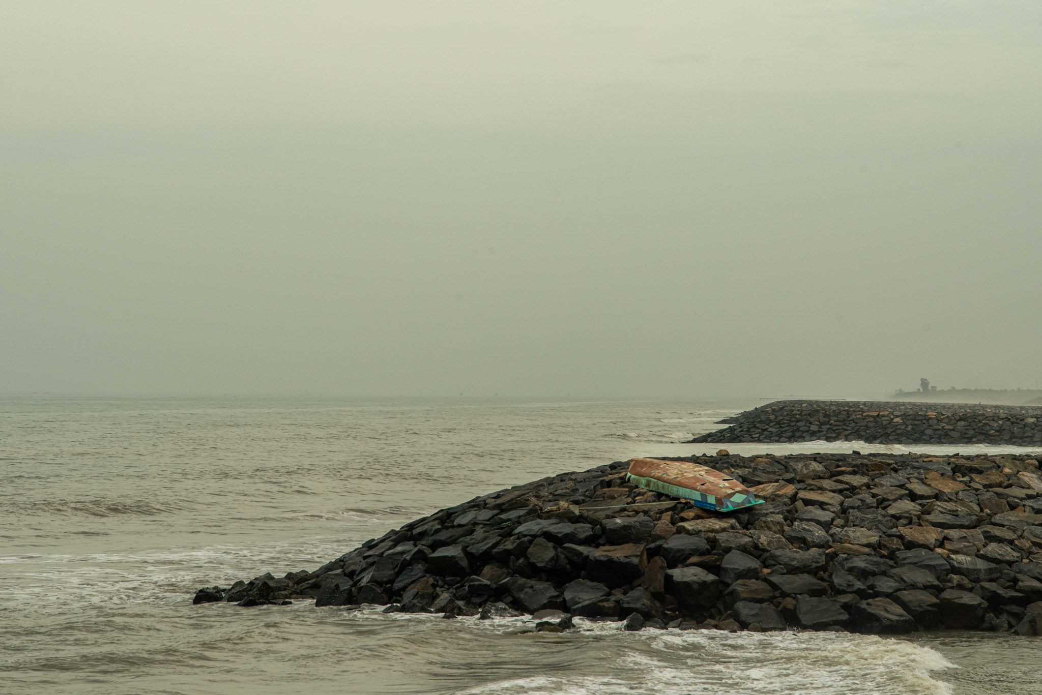 Waves wash against a jetty of dark boulders under a white sky.
