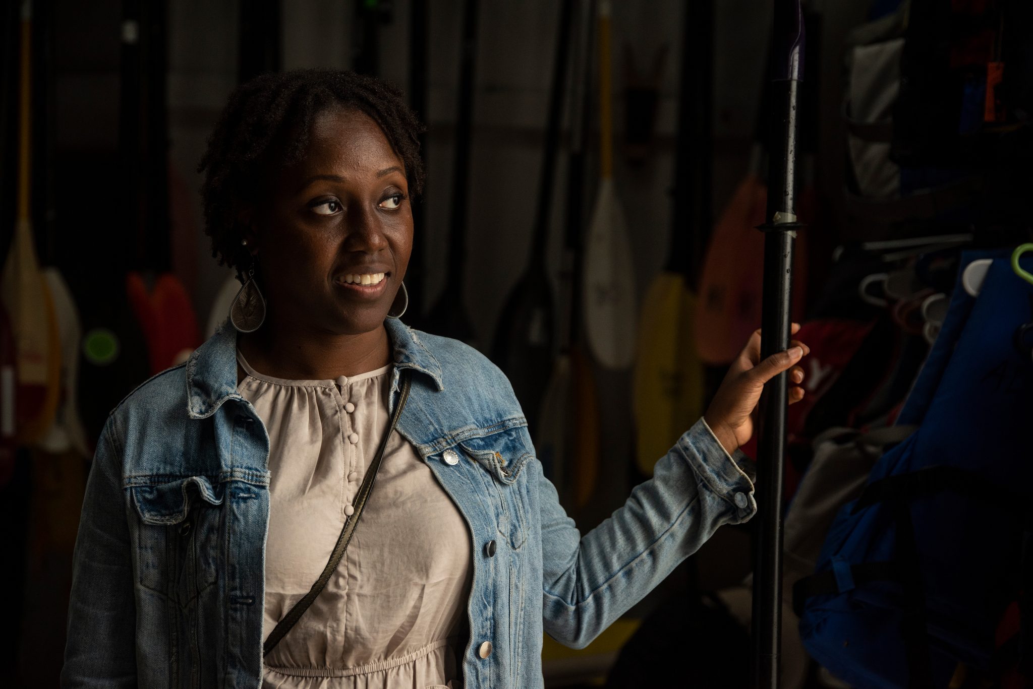 In the dim boathouse a woman in a denim jacket smiles beside the paddle racks.