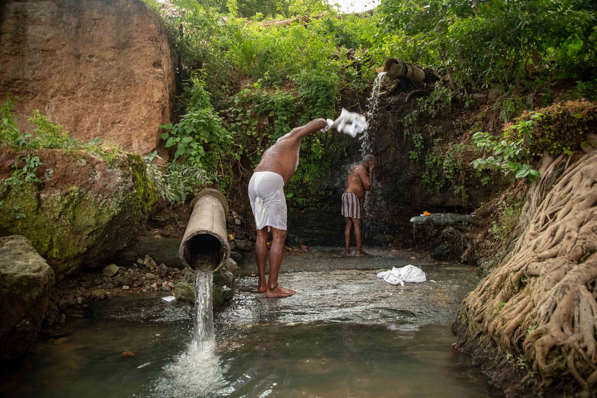 A man washes beneath water spilling from the rocks, a pipe pouring into the pool below.