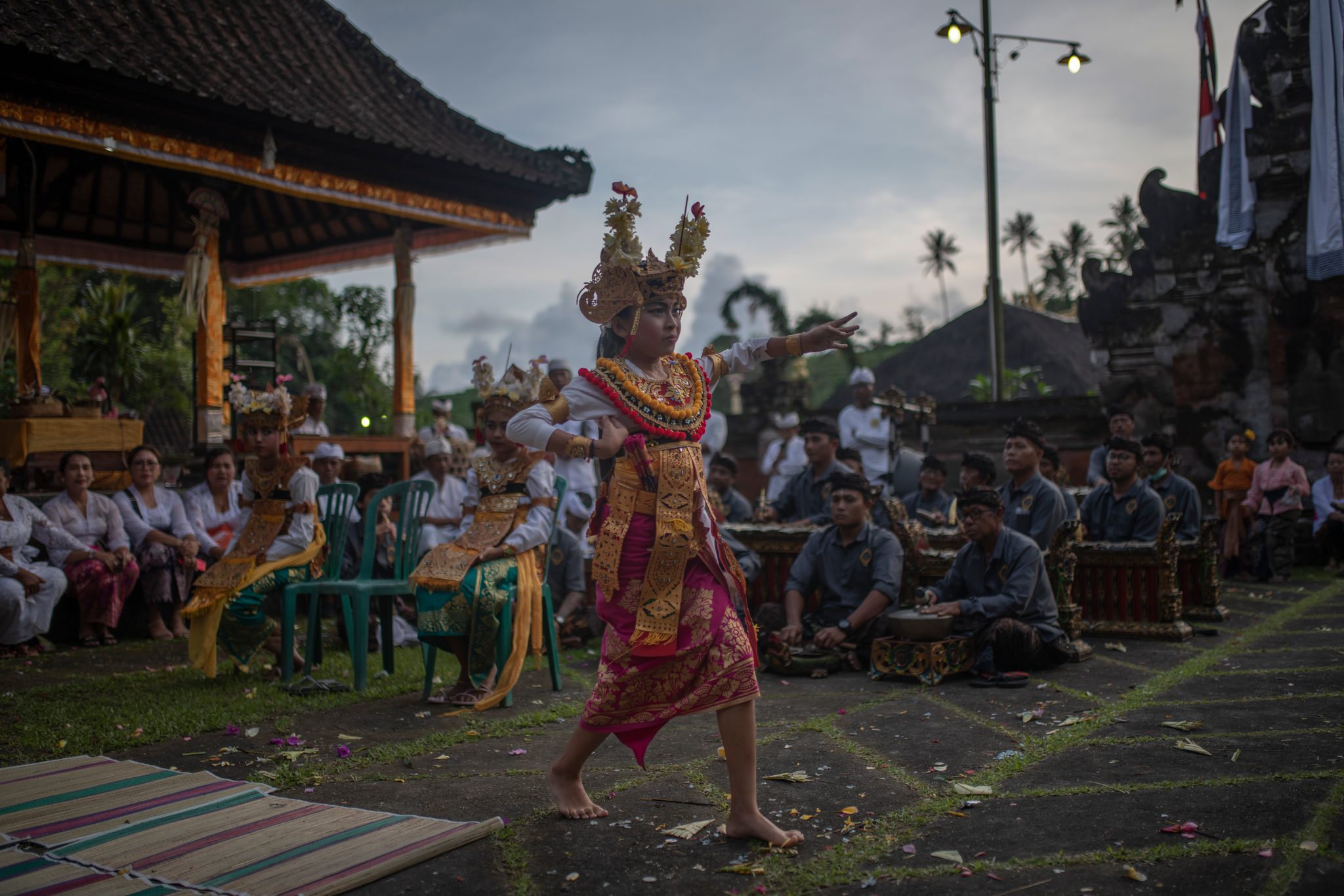 A dancer in gold and a tall crown steps across the grass as the crowd watches at dusk.