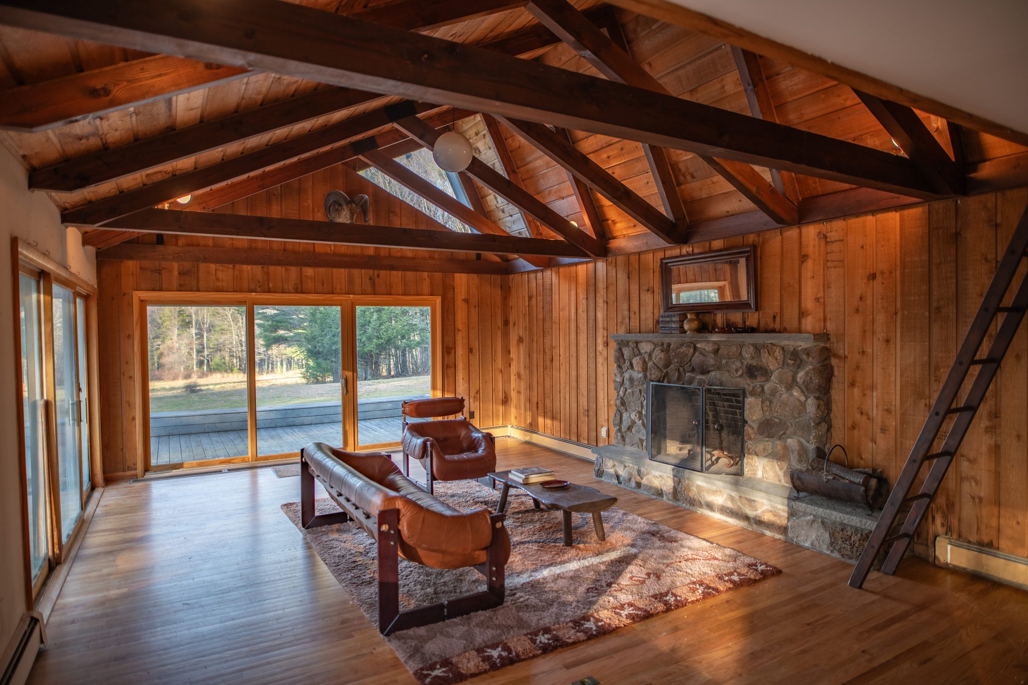 December sun reaches deep into a mid-century great room of wood paneling, exposed beams and a fieldstone fireplace.