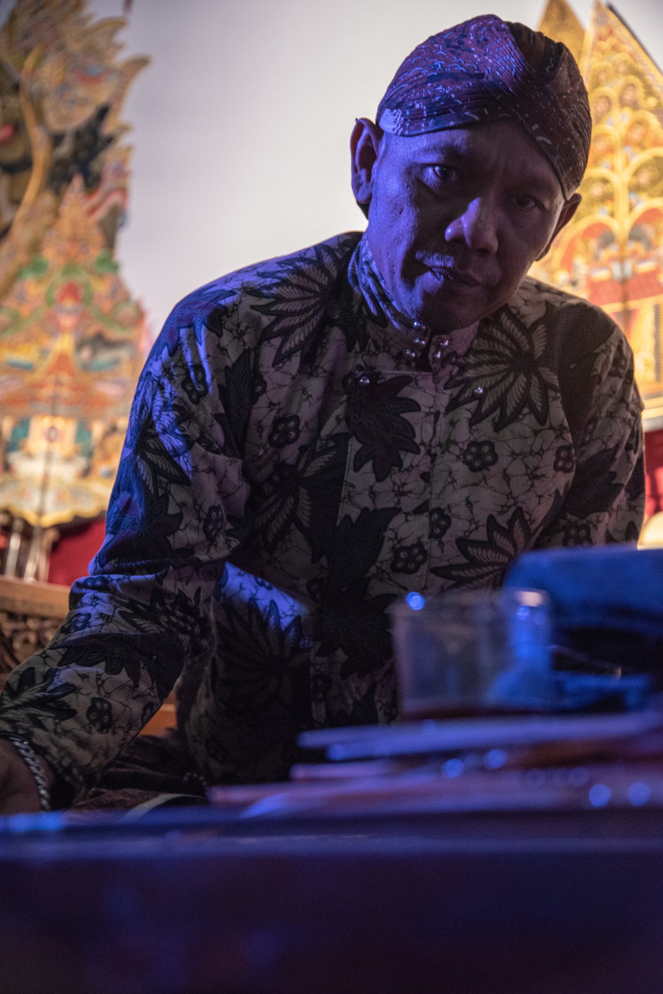 A man in a batik headcloth leans over the wooden chest, the puppets standing at his back.