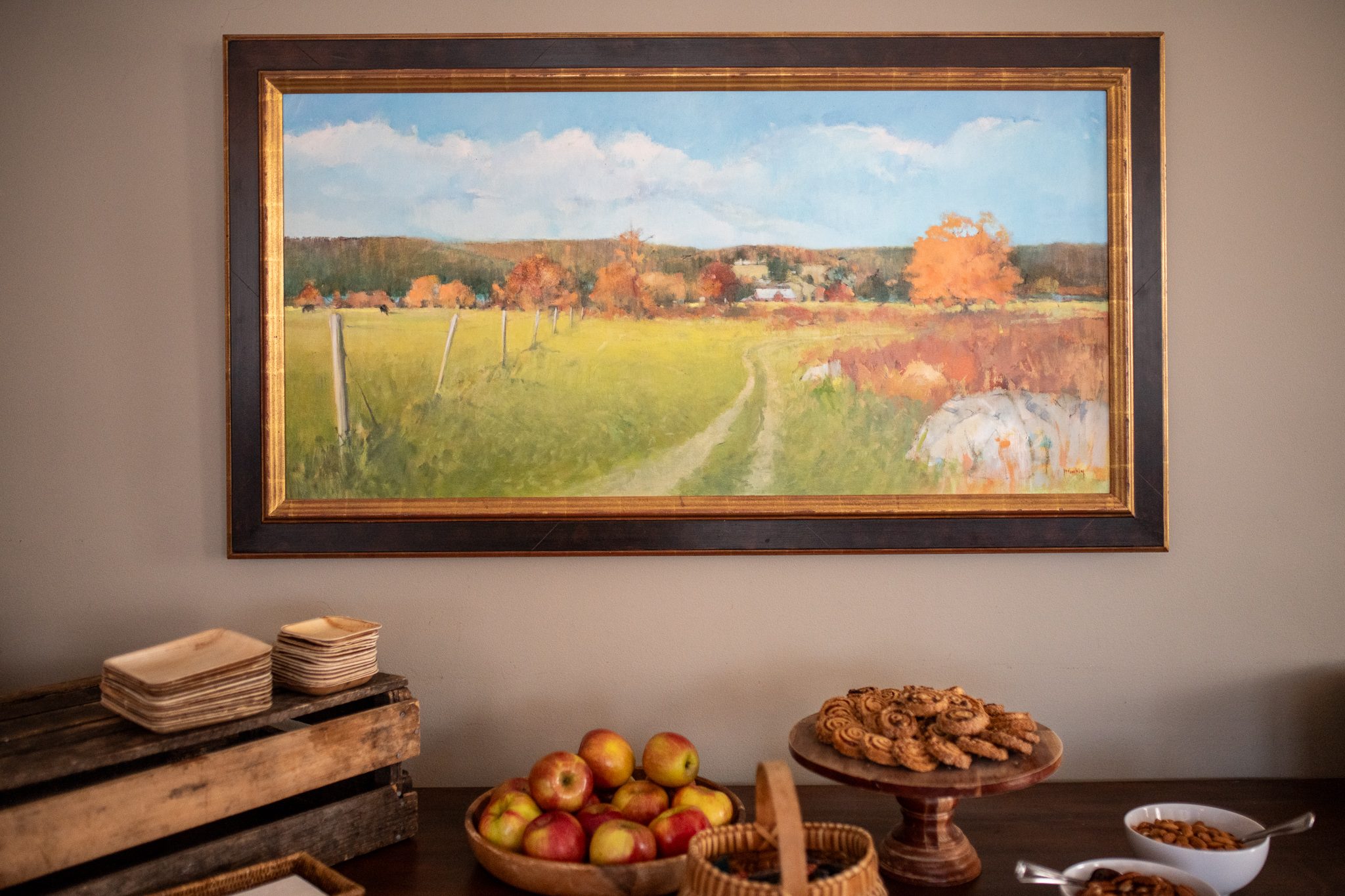 Apples and pastries set out beneath a painted autumn field.
