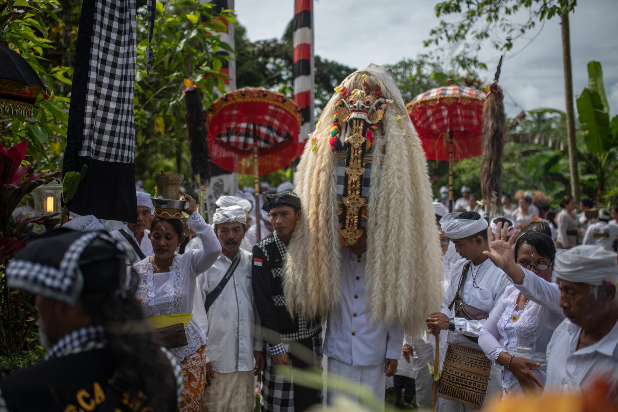 The masked figure moves through the crowd beneath red parasols, its white mane nearly touching the ground.