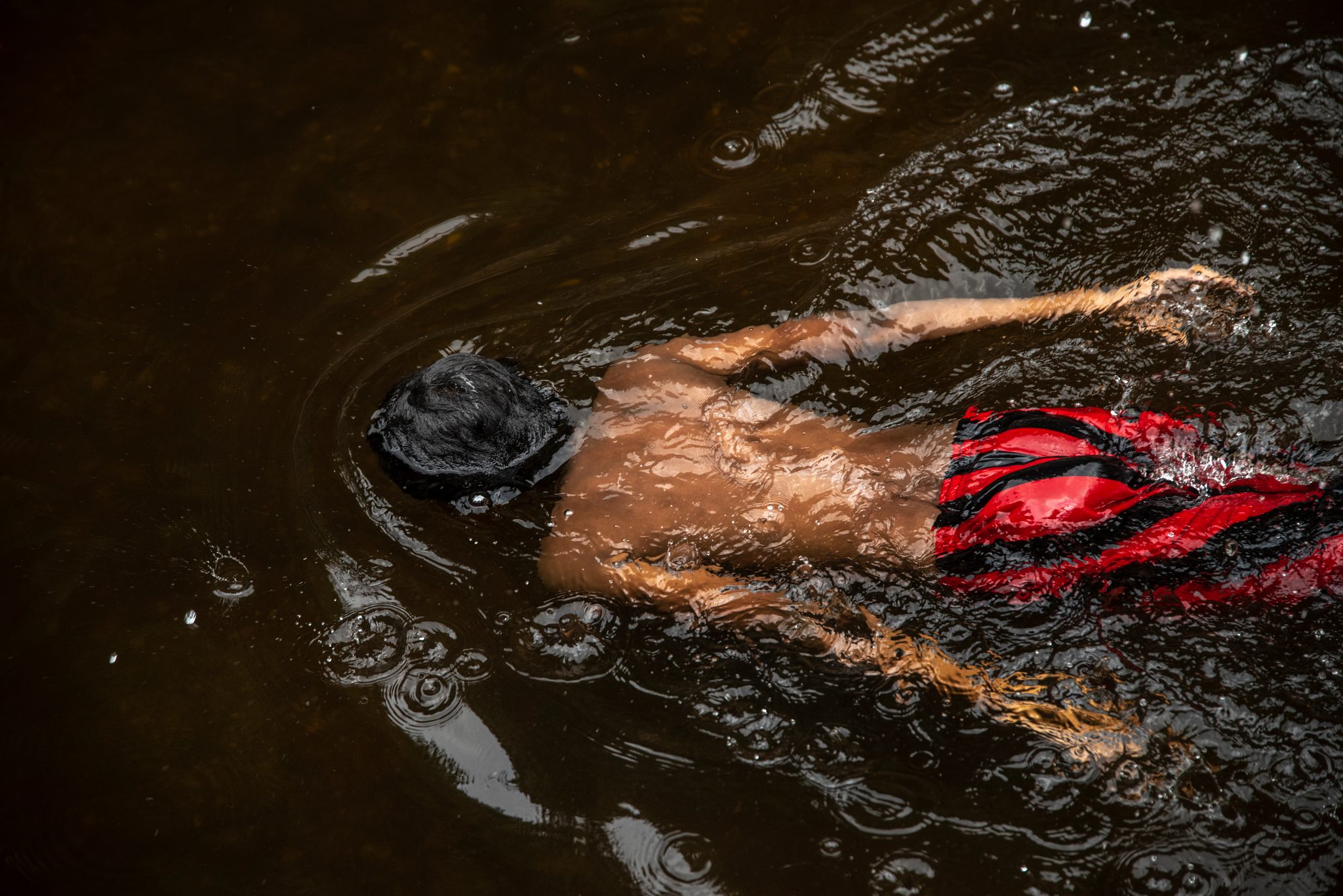 A boy swims face down through the dark water, his red shorts the brightest thing in the river.