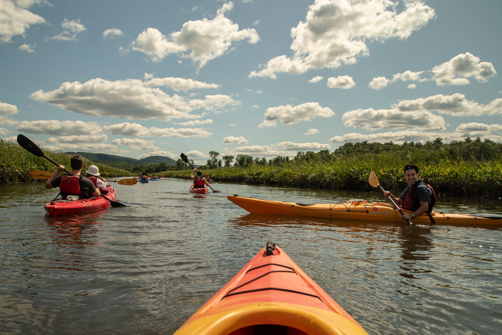From the bow of a kayak, the group paddles up the marsh under summer clouds.